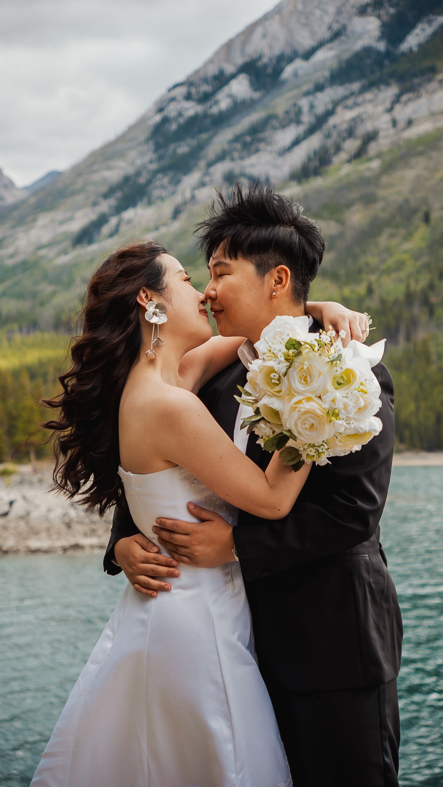A couple shares a romantic moment at their mountain lakeside wedding with a white rose bouquet and scenic backdrop.