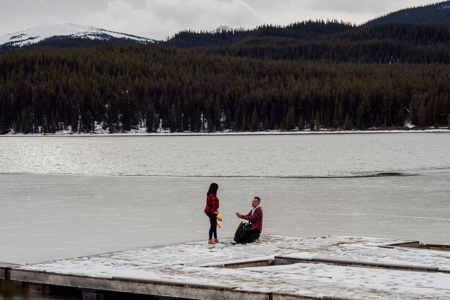 A winter proposal scene on a frozen lake surrounded by snow-capped mountains.