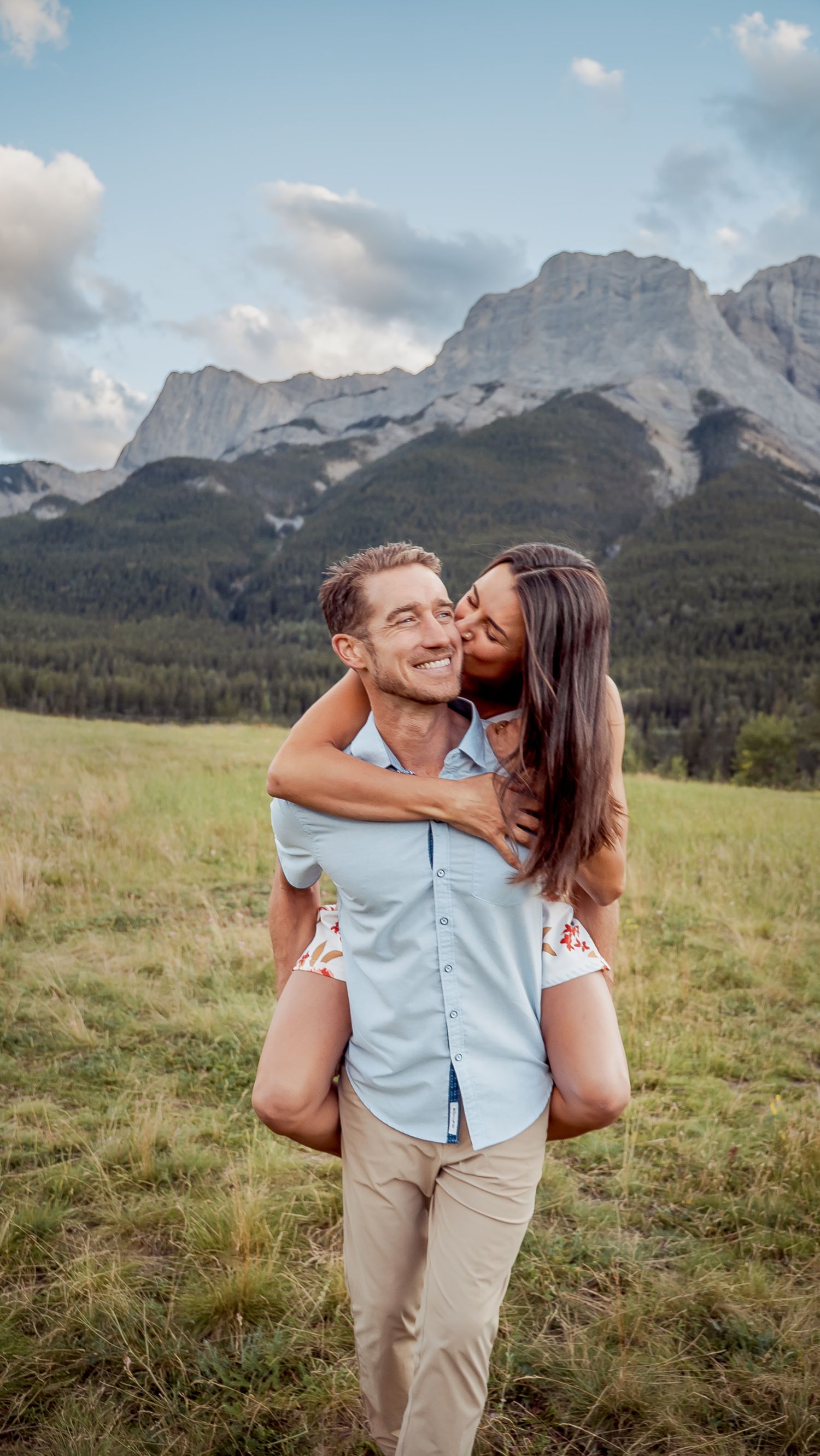 A couple shares a playful moment in a mountain meadow with dramatic Rocky Mountain peaks in the background.