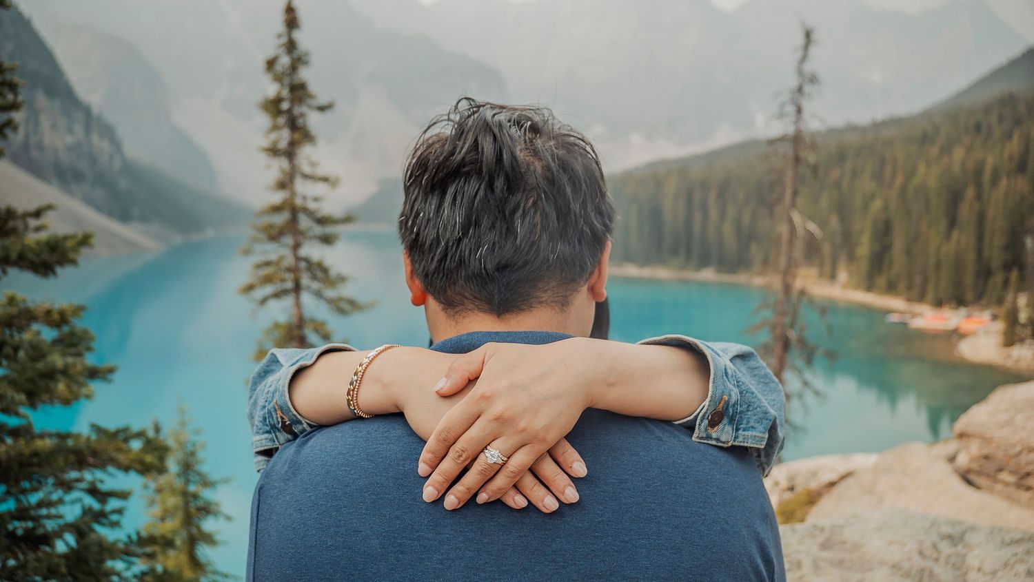 A couple embraces while overlooking a scenic mountain lake surrounded by evergreen forests and rocky peaks.