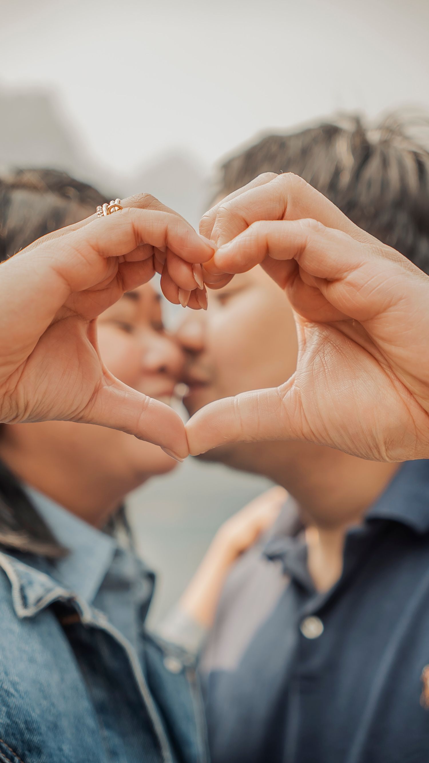 Two hands forming a heart shape while a couple shares an intimate moment in denim clothing.