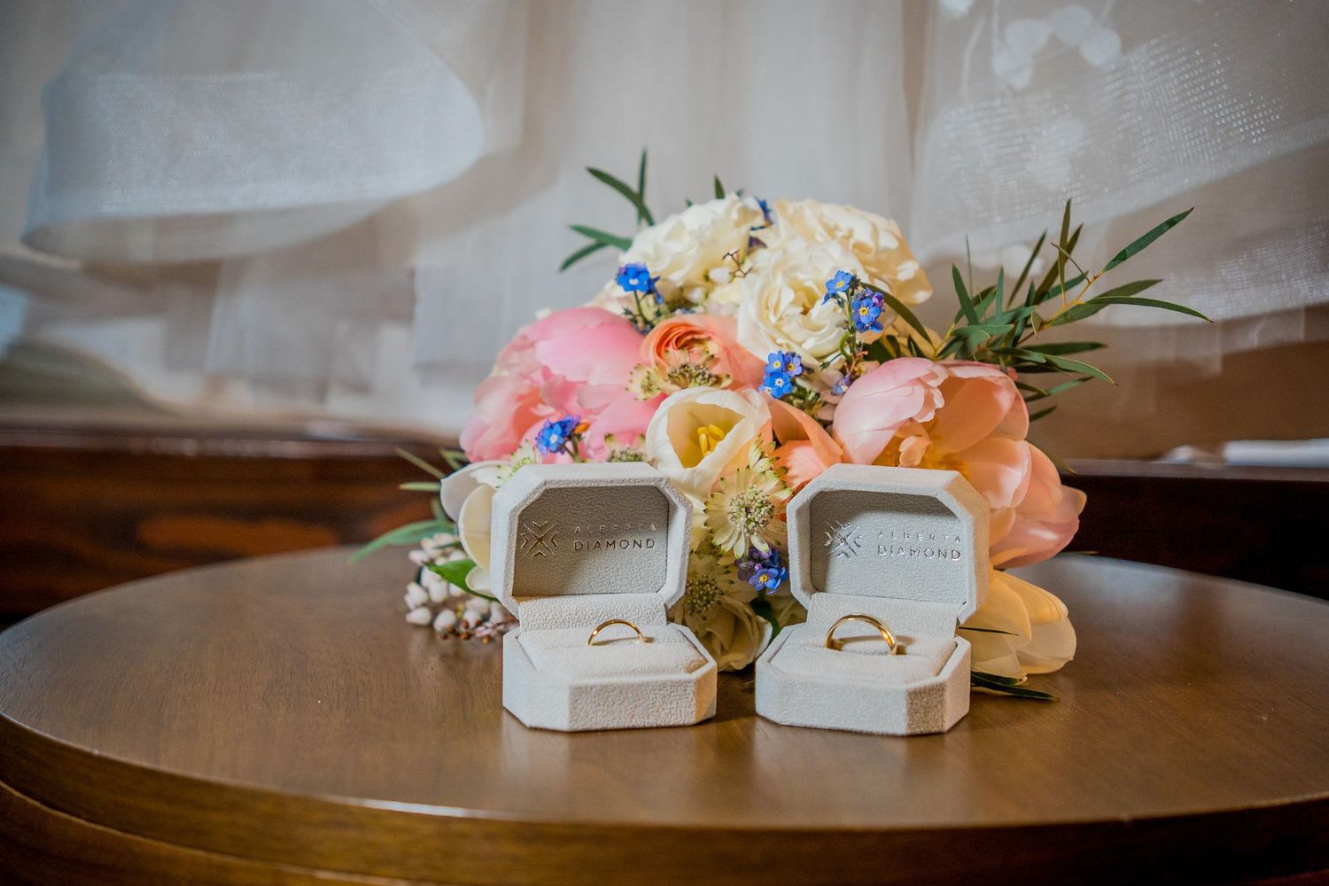 Two gold wedding rings in white velvet boxes displayed on a wooden table beside a colorful bridal bouquet.