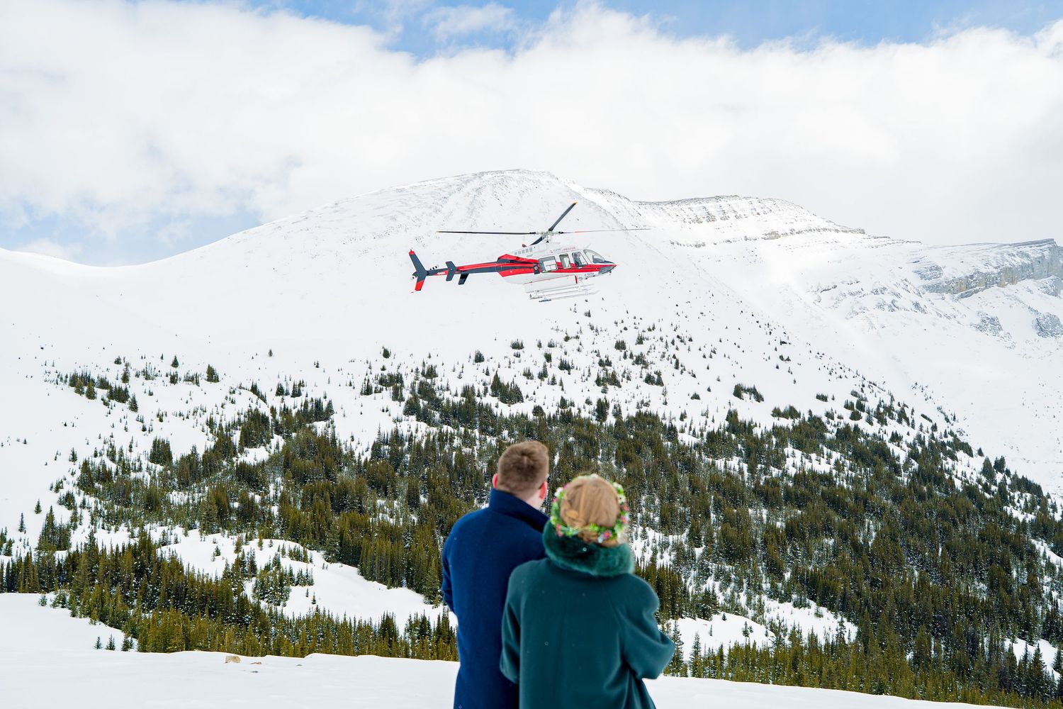 Couple watching a red helicopter fly over a snowy mountain landscape with evergreen trees.