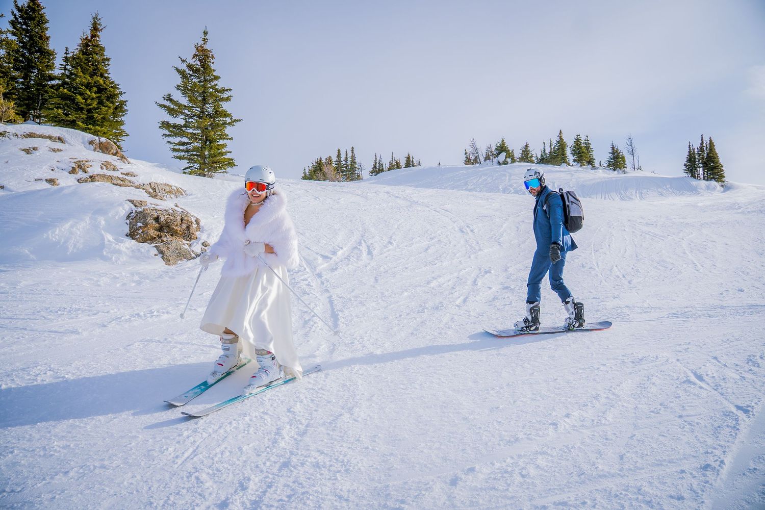 Two people skiing down a snowy slope with evergreen trees in the background.