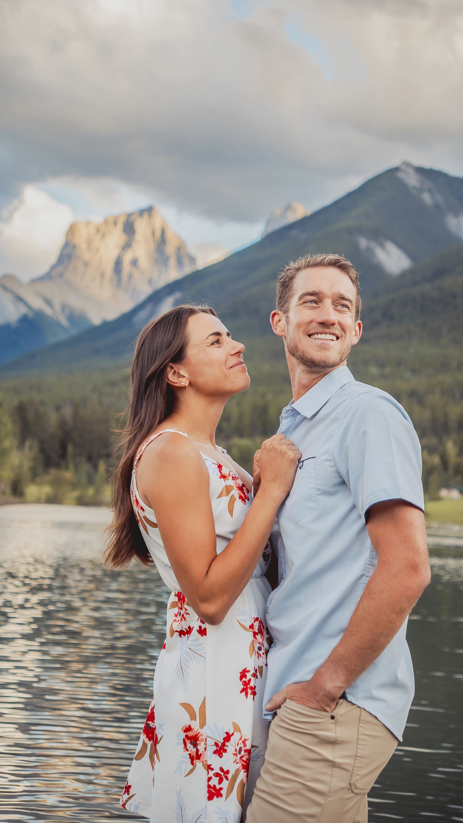 A couple laughs together by a mountain lake at sunset with dramatic peaks rising in the background.