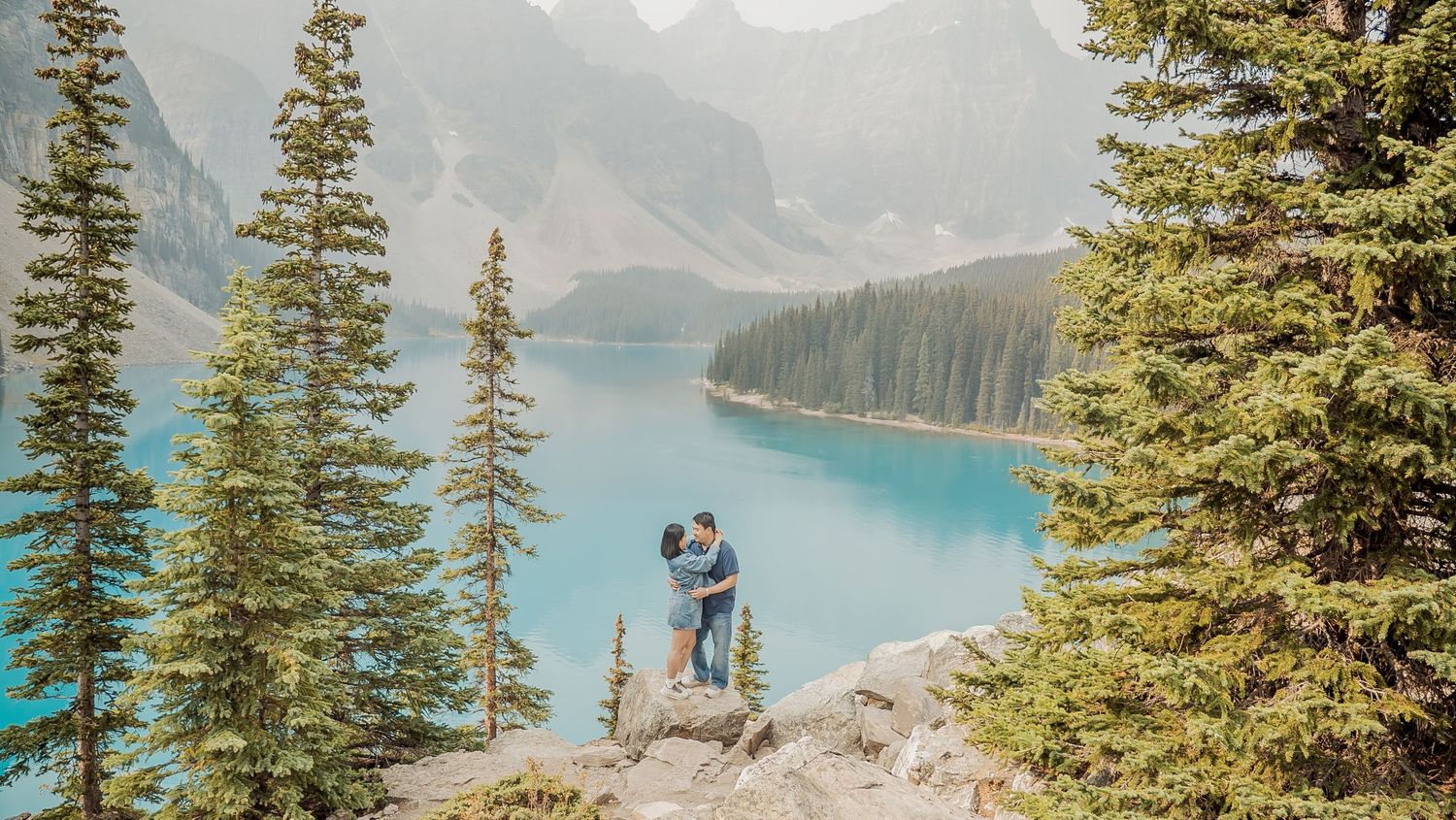 Hikers stand on rocky overlook admiring turquoise Moraine Lake surrounded by evergreen forest and mountain peaks in Banff.