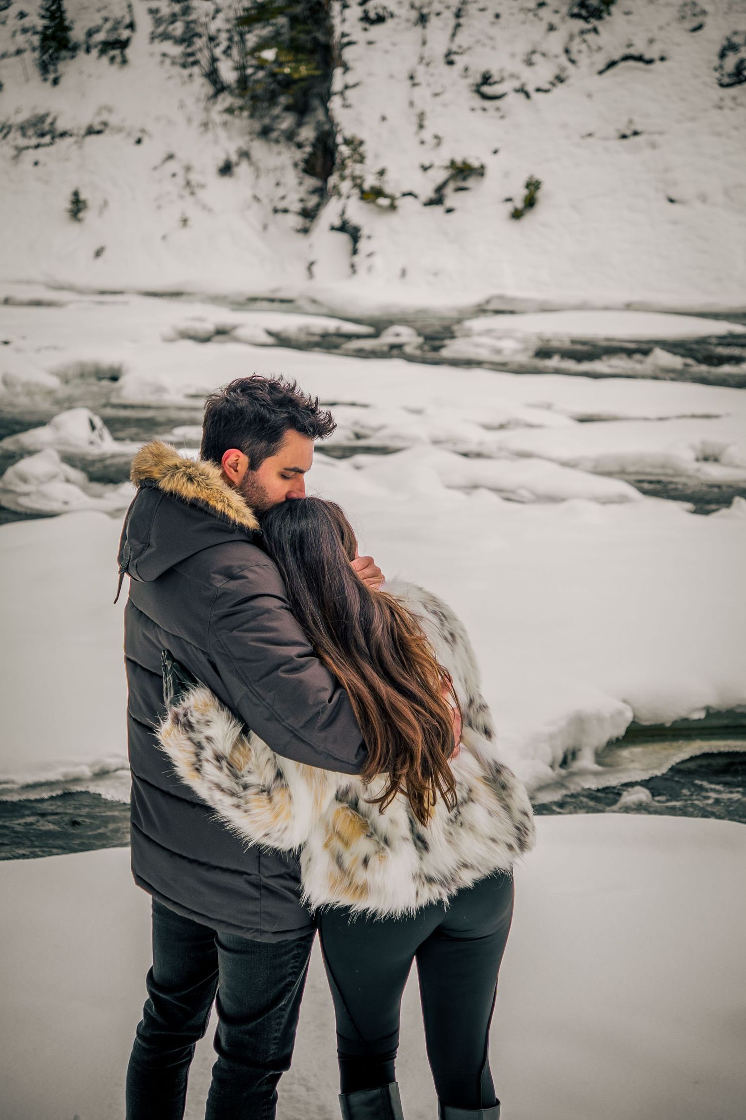 A couple in matching black winter coats shares an intimate moment by a frozen river.