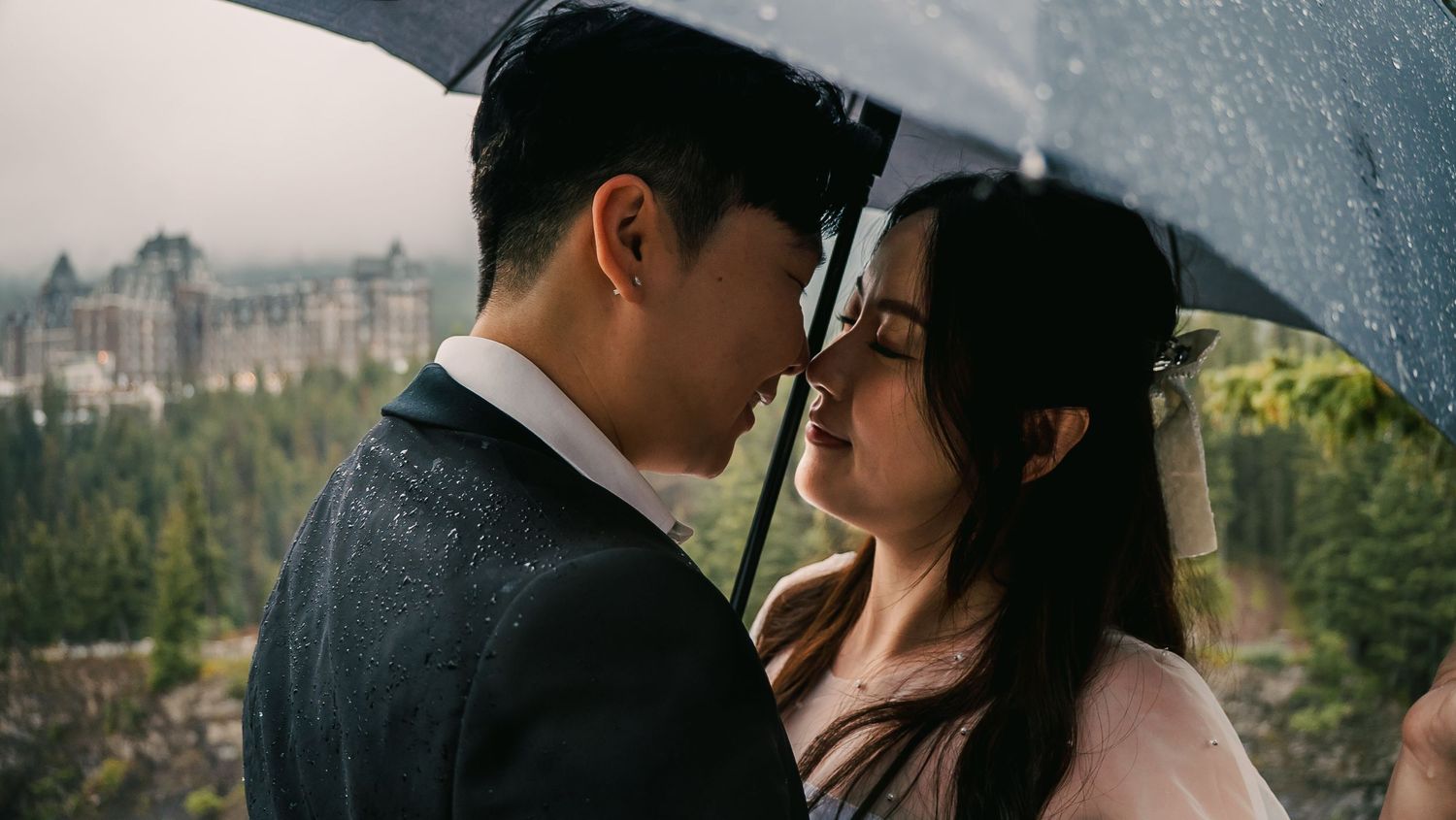 A couple shares an intimate moment under an umbrella in the rain against a scenic mountain landscape.