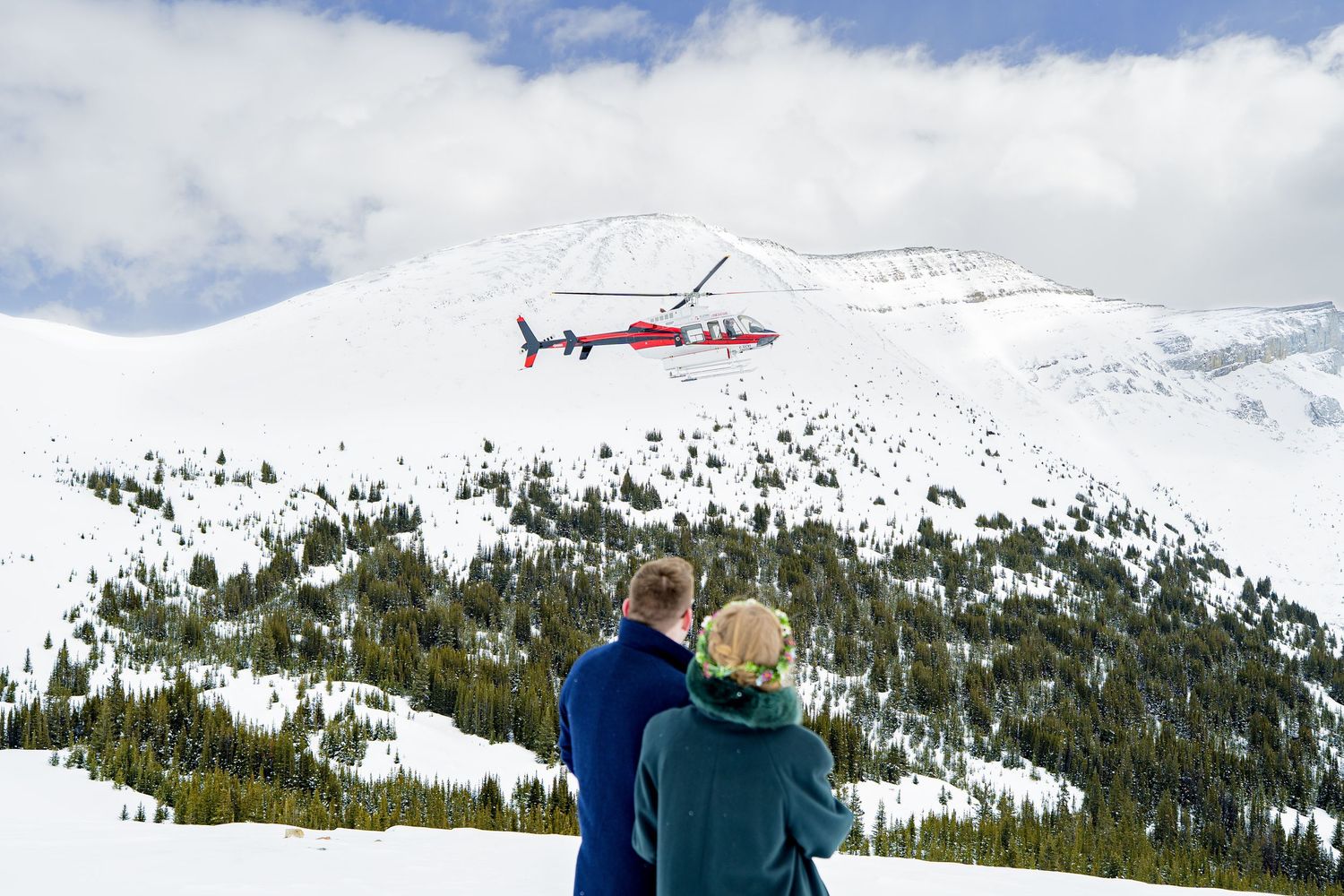 Couple watching a red helicopter fly over a snow-covered mountain under a partly cloudy sky.