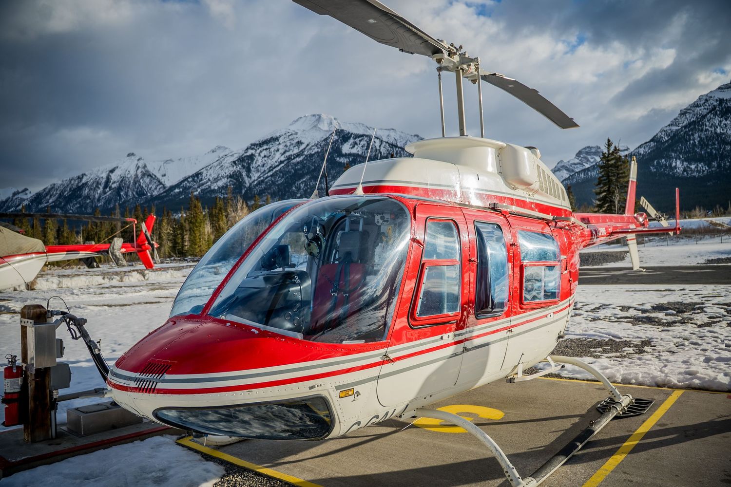 Red and white helicopter parked on snowy helipad with dramatic mountain backdrop in winter landscape.