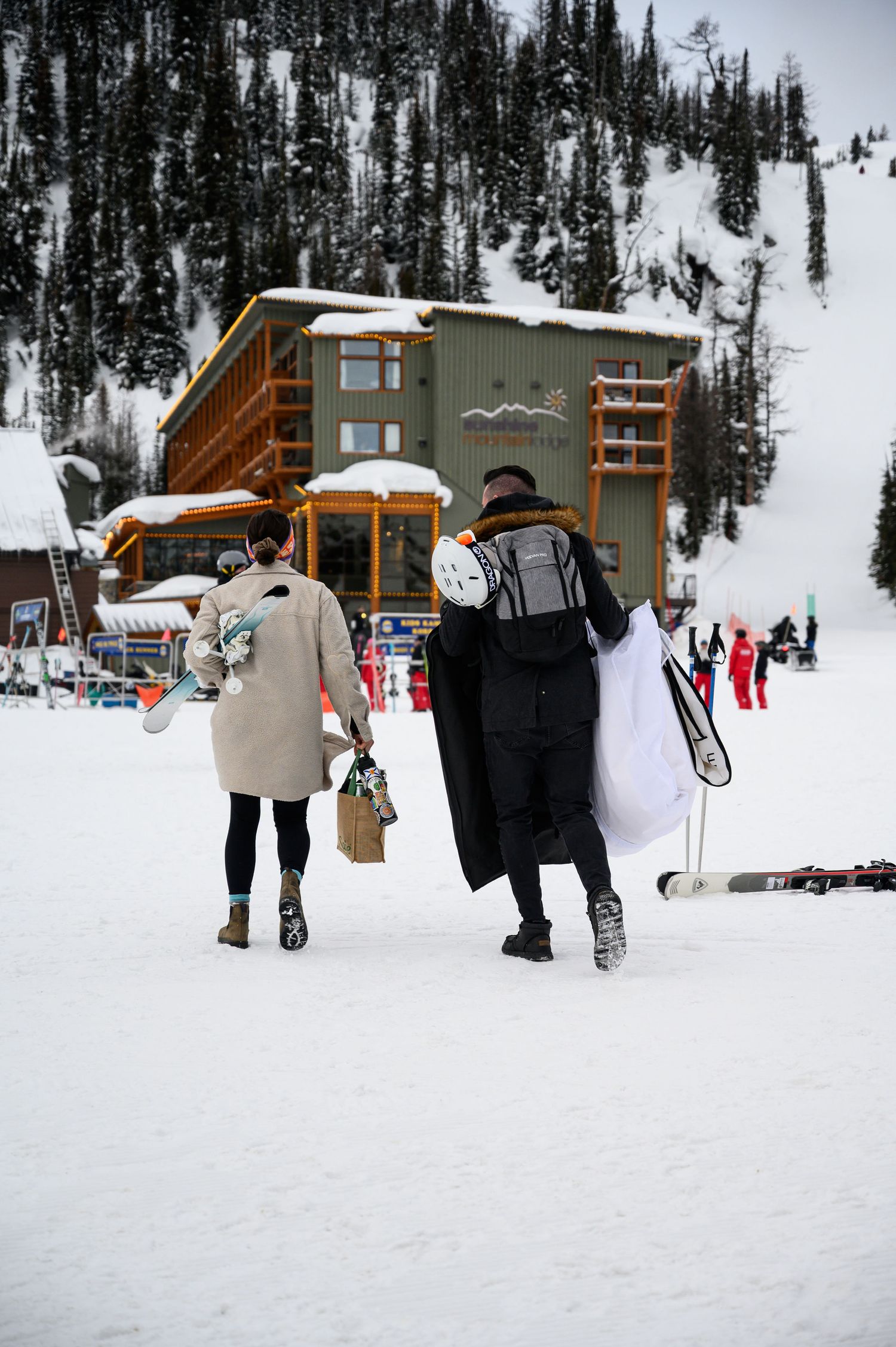 Skiers and snowboarders carry their gear across a snowy base area with wooden lodge buildings in the background.