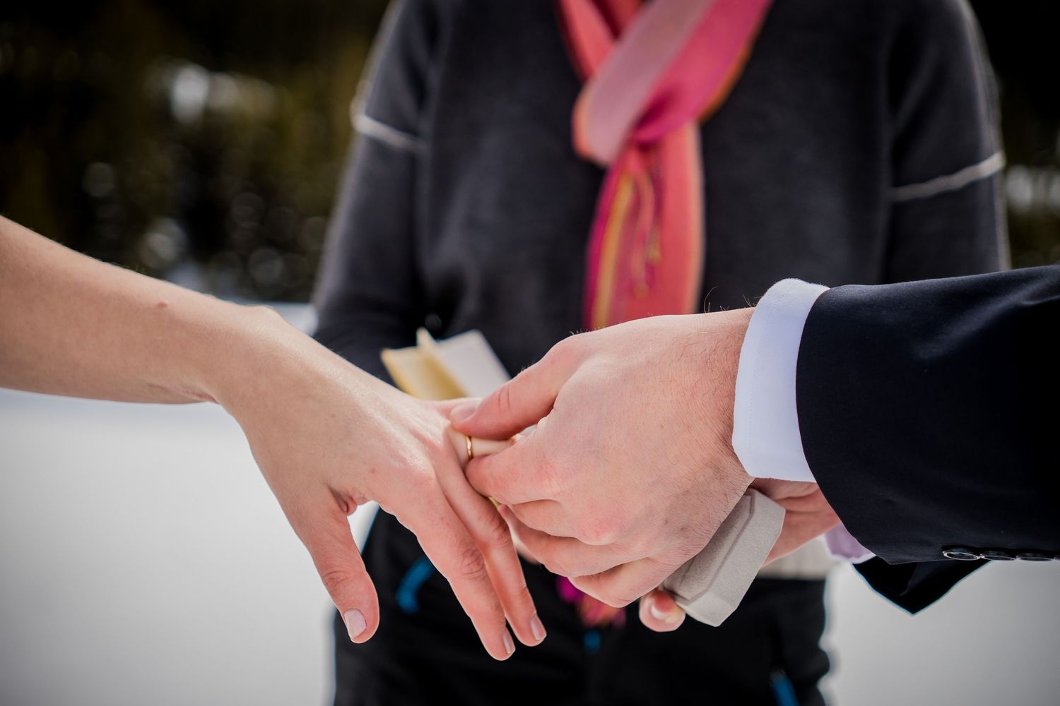 Close-up of a couple exchanging rings during a wedding ceremony, officiant in pink scarf visible behind them.
