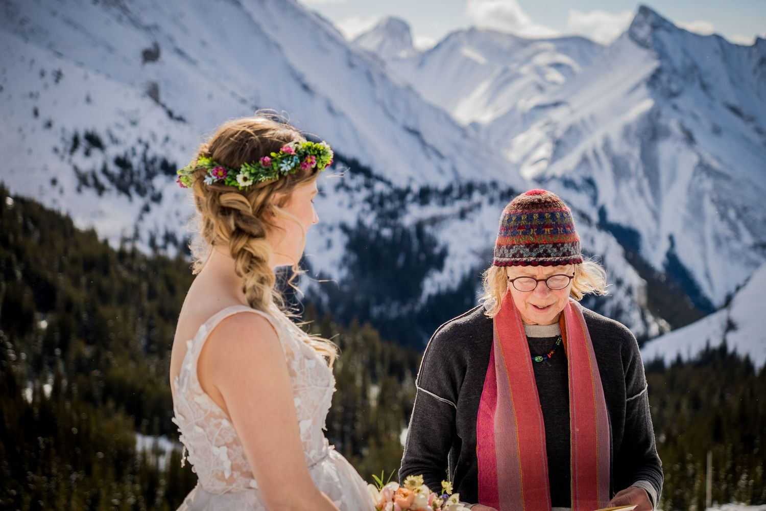 Bride with floral crown exchanges vows with officiant against snowy mountain backdrop at winter wedding.