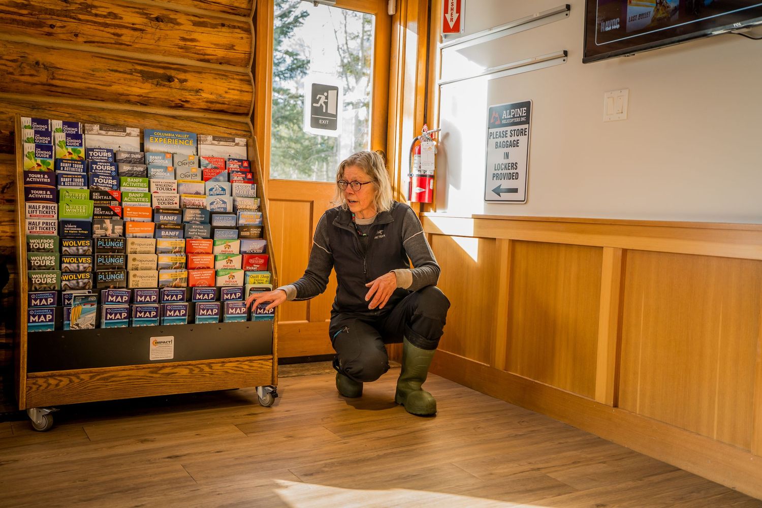 Woman crouching near a brochure display rack inside a wooden visitor center or lodge.