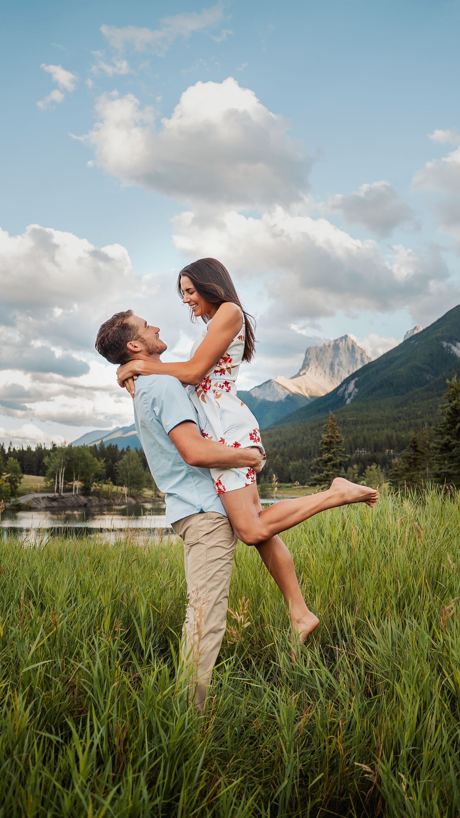Couple shares joyful moment in mountain field with dramatic cloudy sky and peaks in background.