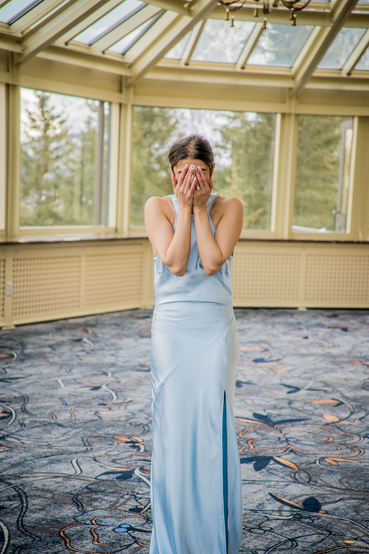A person in a light blue dress stands in an abandoned room with glass windows and scattered debris on the floor.