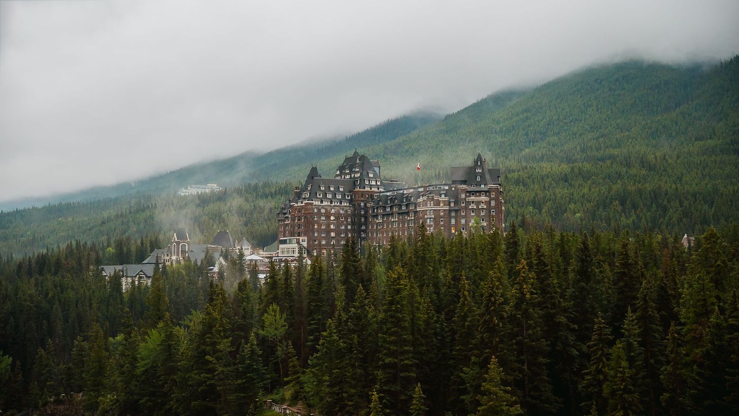 A panoramic mountain landscape with ski resort buildings nestled among evergreen forests and snow-capped peaks.