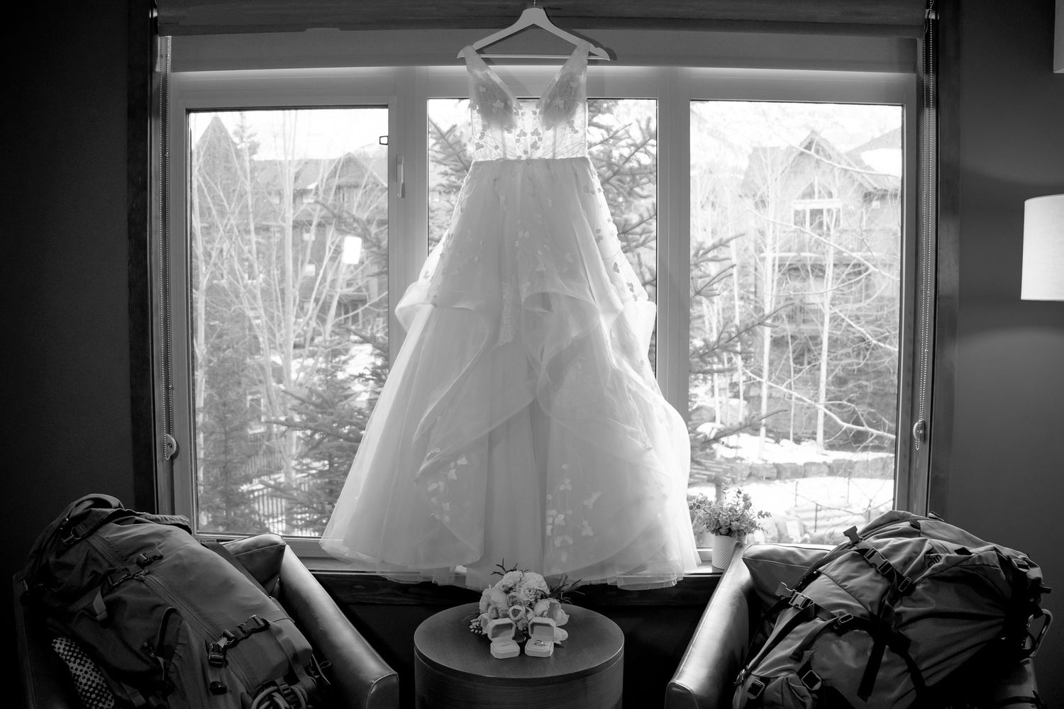 Black and white photo of an elegant wedding dress hanging in a window with floral bouquet below.