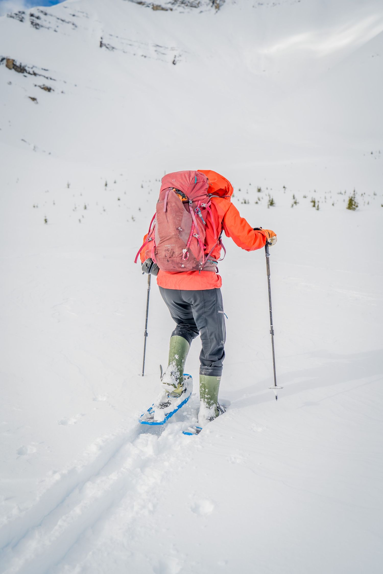 A hiker in a red jacket with a backpack uses ski poles while snowshoeing up a snowy mountain slope.