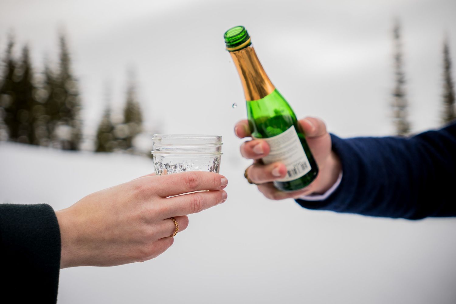 Two hands clinking a green champagne bottle and glass outdoors in a snowy winter landscape.