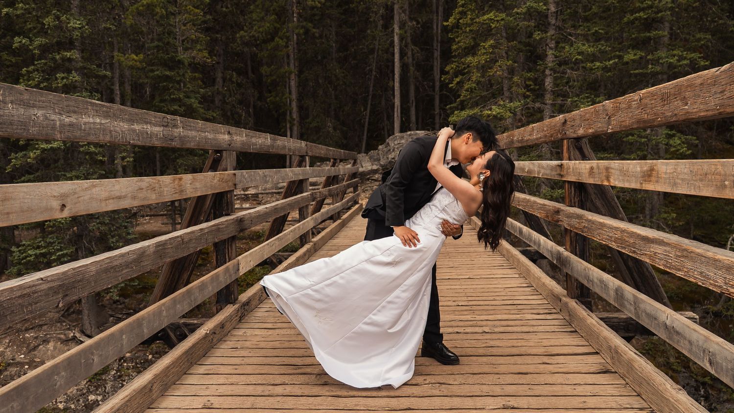 A couple in formal wedding attire embraces on a rustic wooden bridge surrounded by evergreen forest.