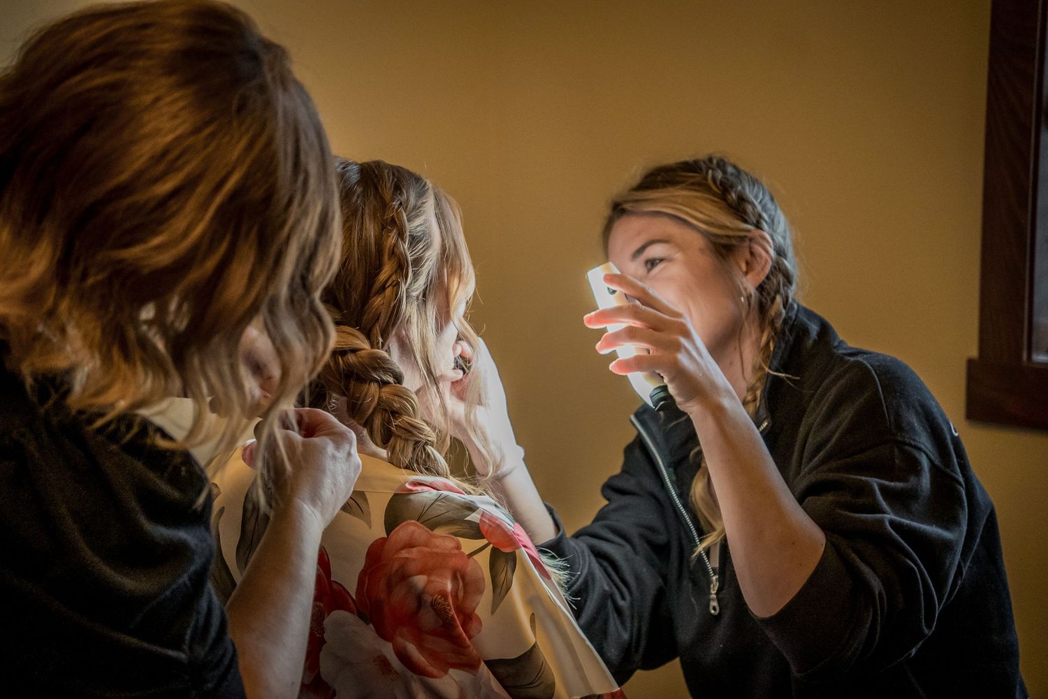 A makeup artist applies cosmetics to a bride while another bridesmaid assists during wedding preparations indoors.