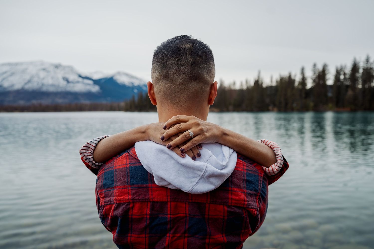 A couple in plaid flannel embraces near a serene mountain lake.