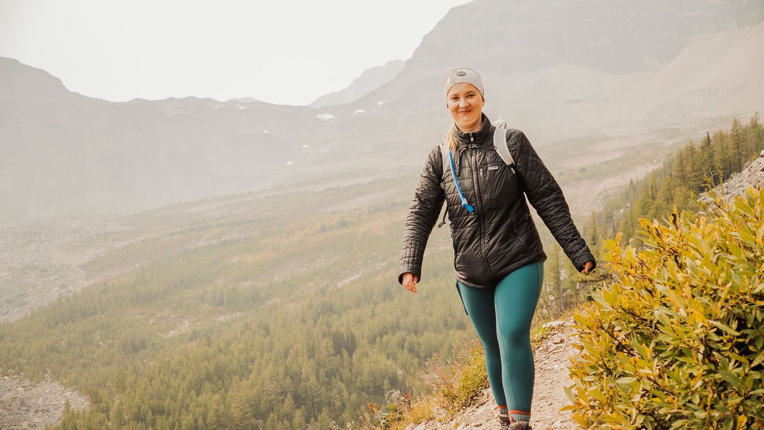 Hiker in athletic wear walks along mountain trail with scenic valley views and yellow wildflowers in foreground.