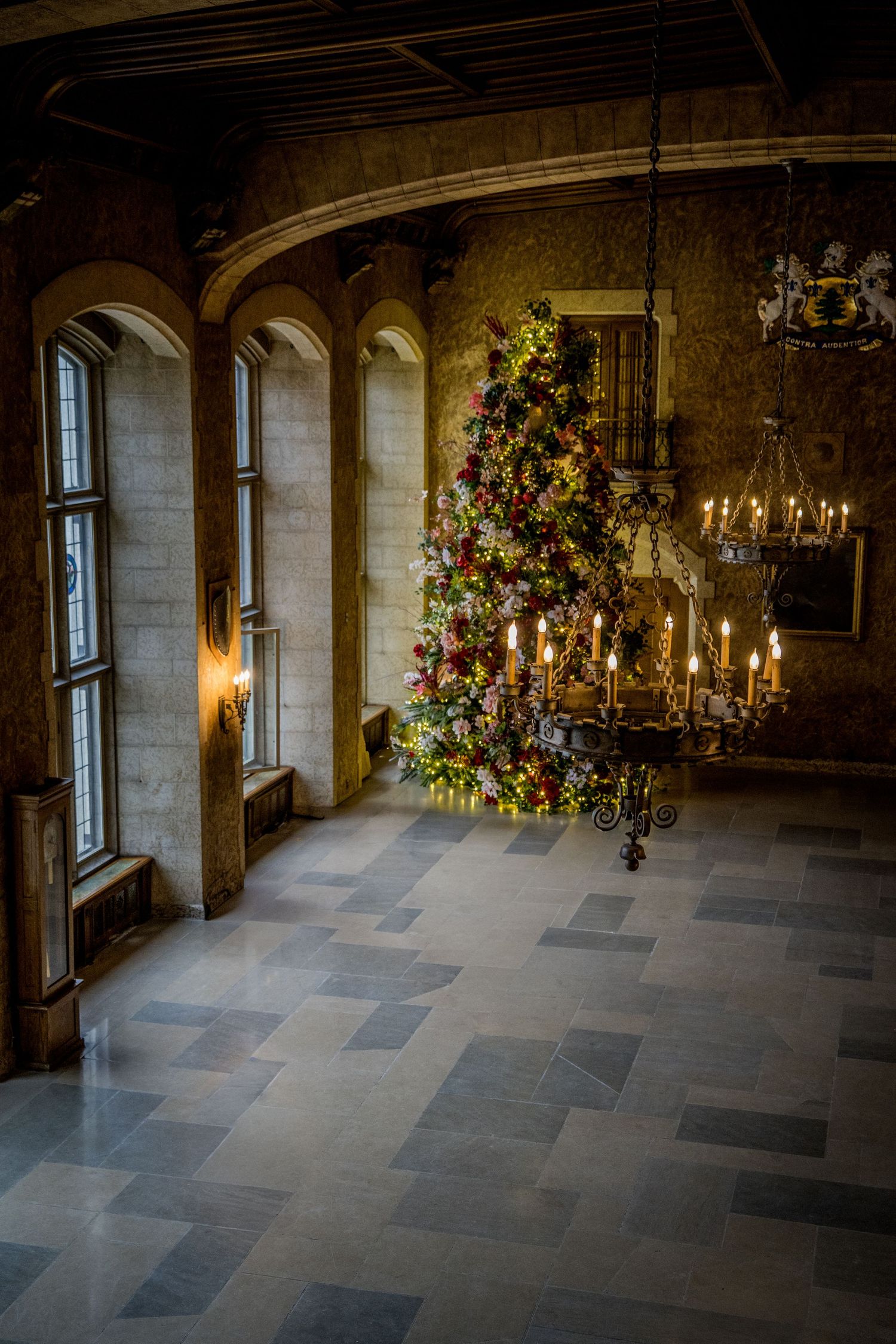 Elegant stone hall with chandeliers and holiday decorations in warm lighting.
