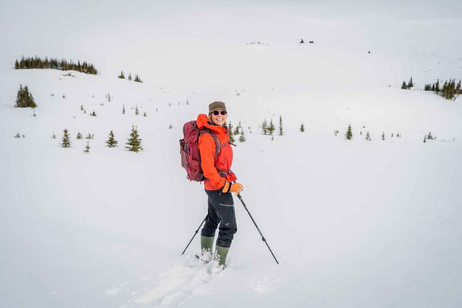 A hiker in an orange jacket with a red backpack and ski poles stands in a snowy mountain landscape with evergreen trees.