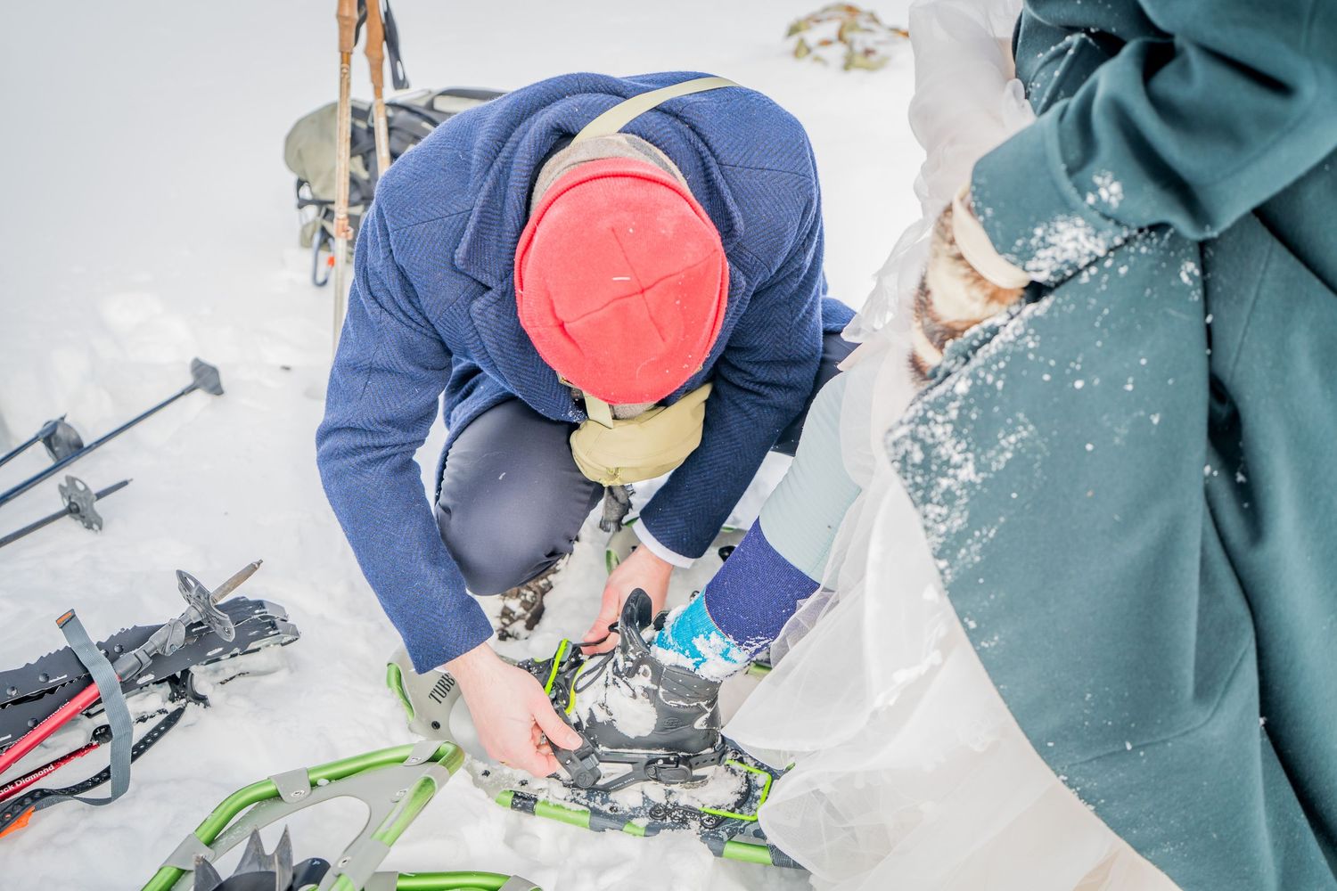 Person in red hat adjusting ski bindings on snowy mountain terrain with poles and green snowshoes nearby.