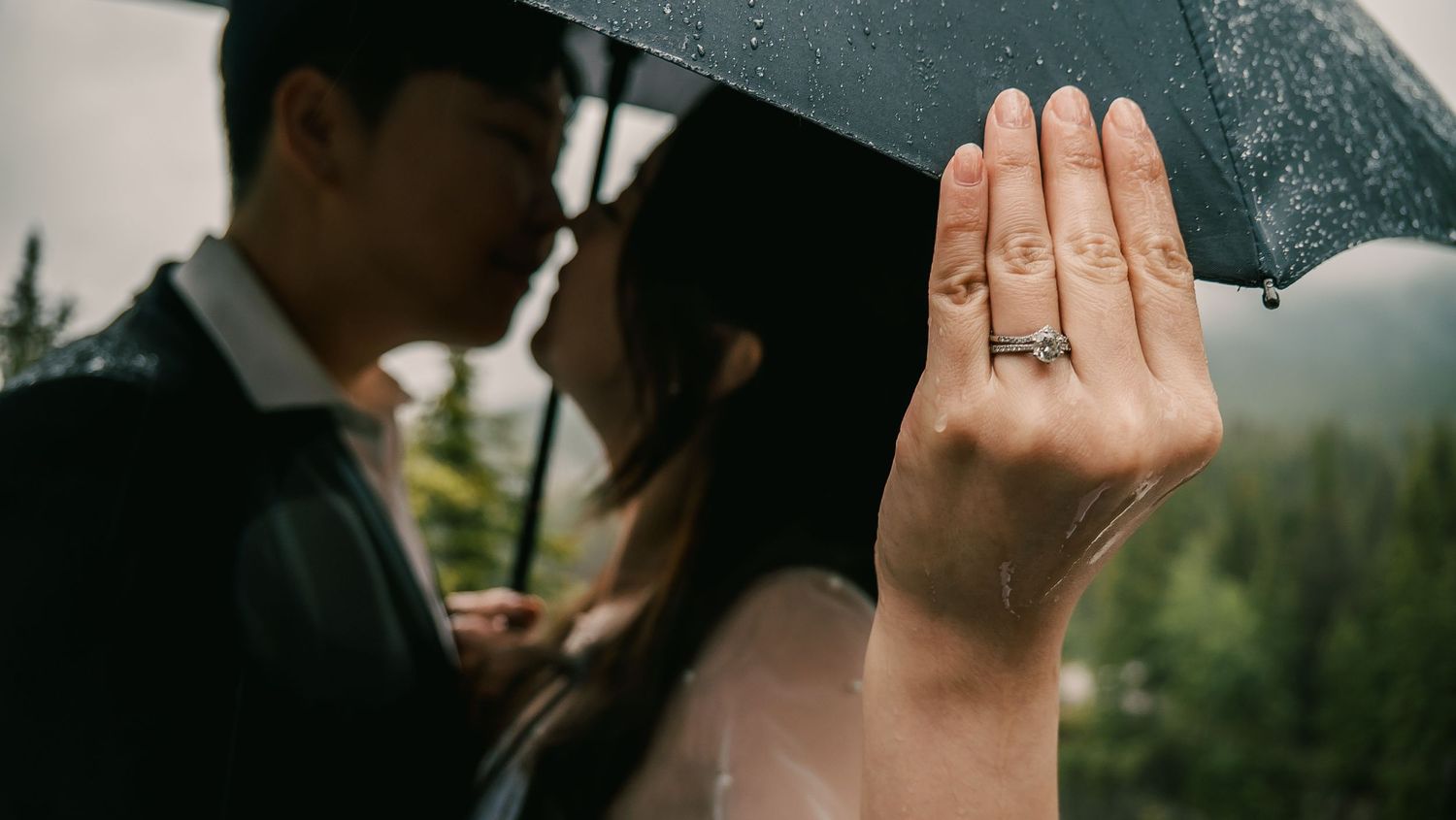A close-up of an engagement ring on a hand while a couple shares an umbrella in the rain.