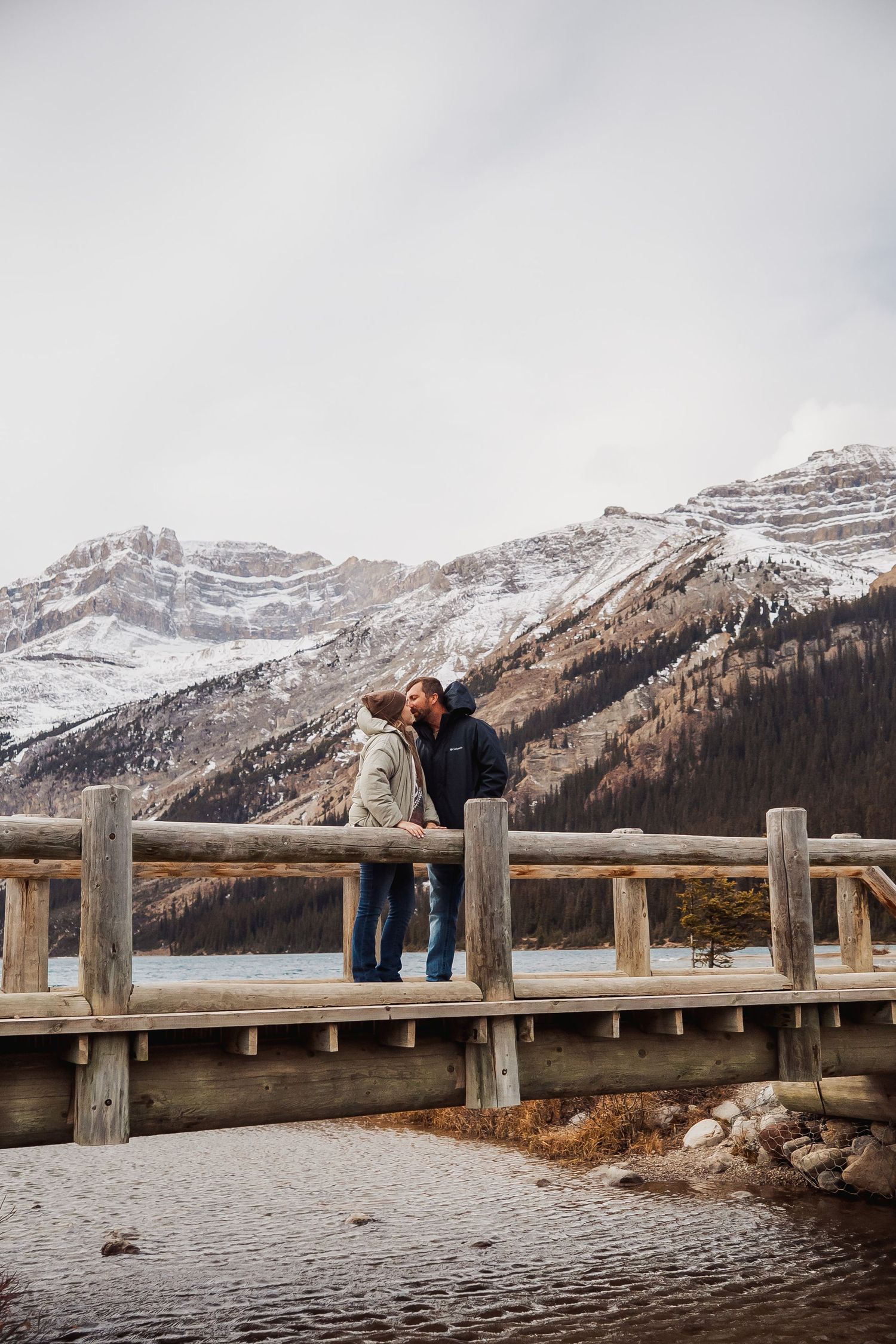 A couple shares a romantic moment on a wooden pier overlooking snow-capped mountains and a serene lake.