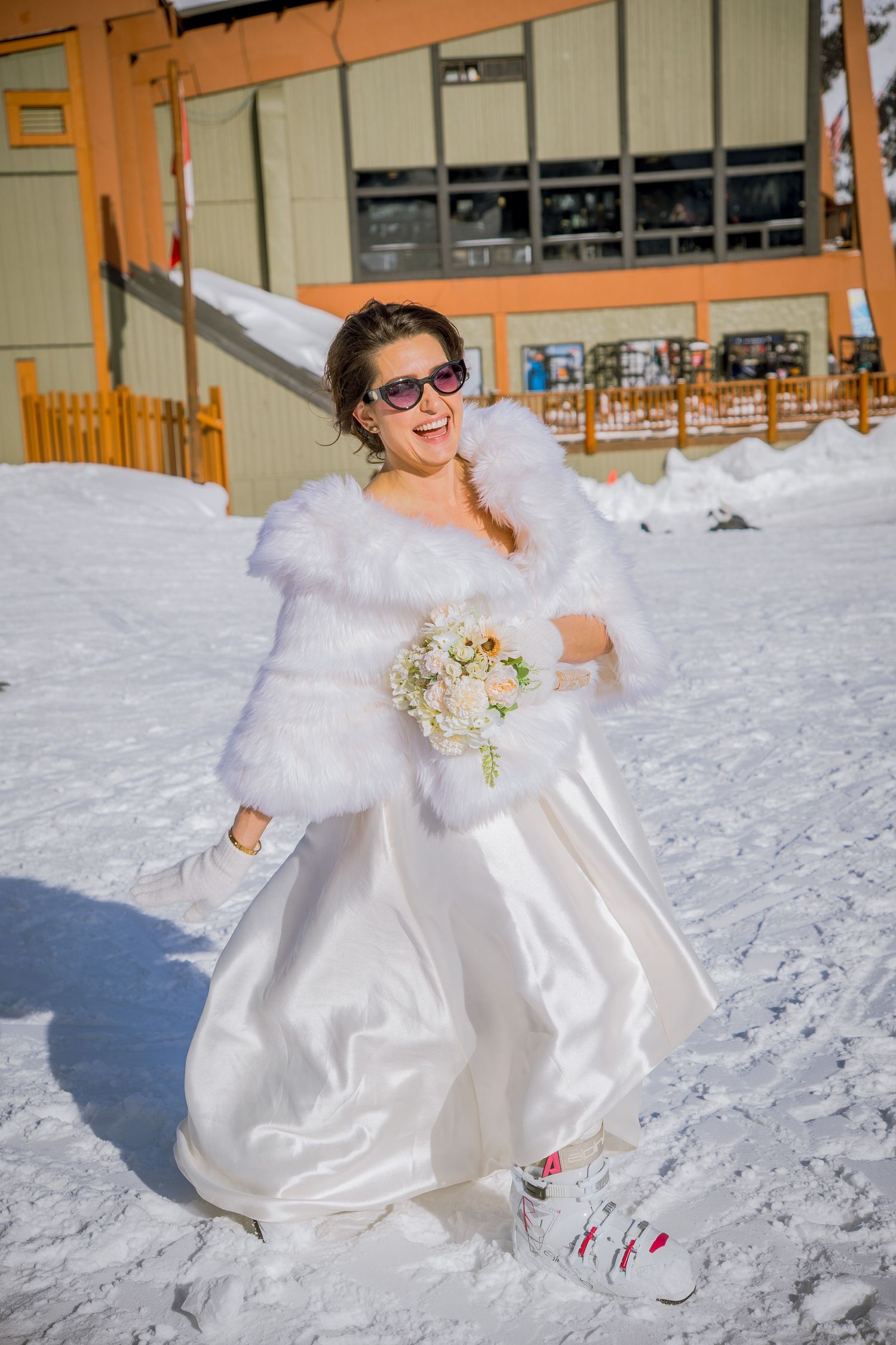 Winter bride in white dress and fur shawl poses playfully in deep snow at a ski resort.