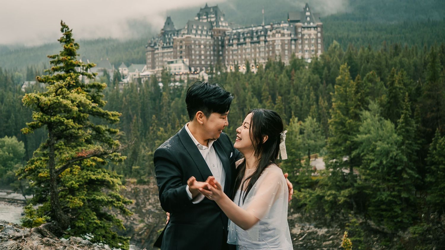 A couple embraces in front of the majestic Banff Springs Hotel surrounded by mountain peaks and evergreen forest.
