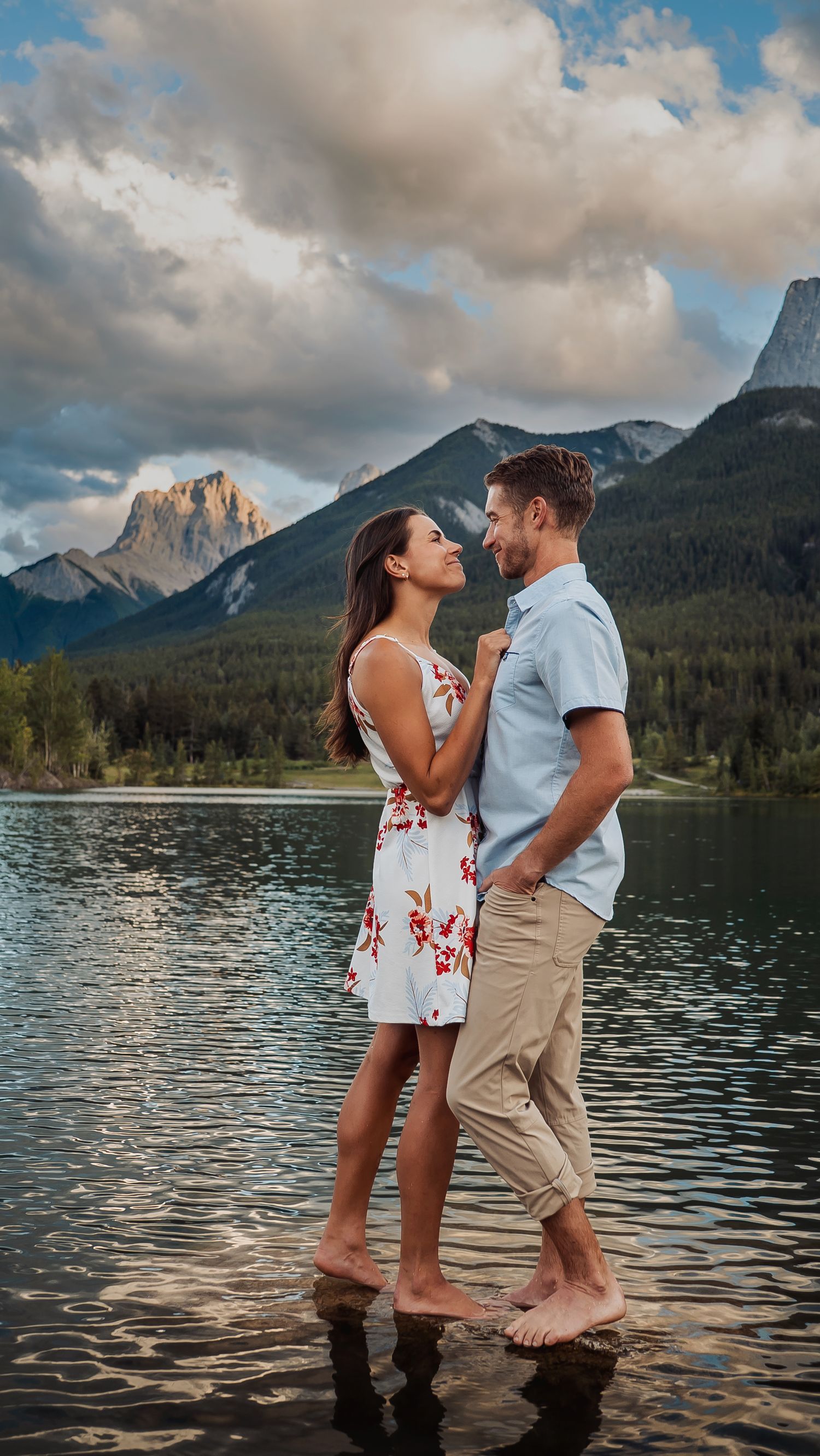 Couple stands intimately in shallow lake water with mountain landscape and dramatic clouds behind them.