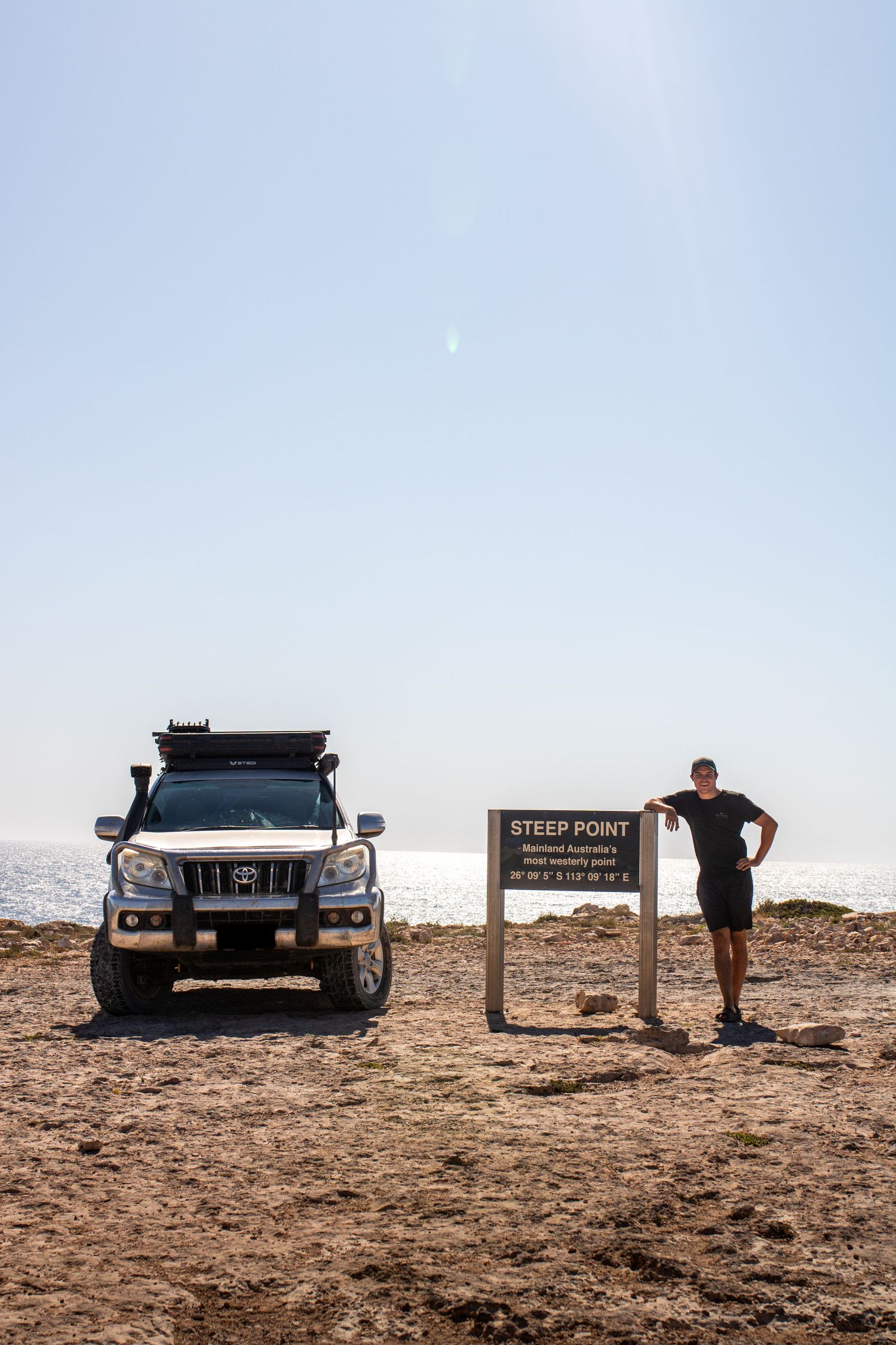 Man standing beside Steep Point sign next to Toyota Land Cruiser on rocky terrain, Australia's westernmost point.