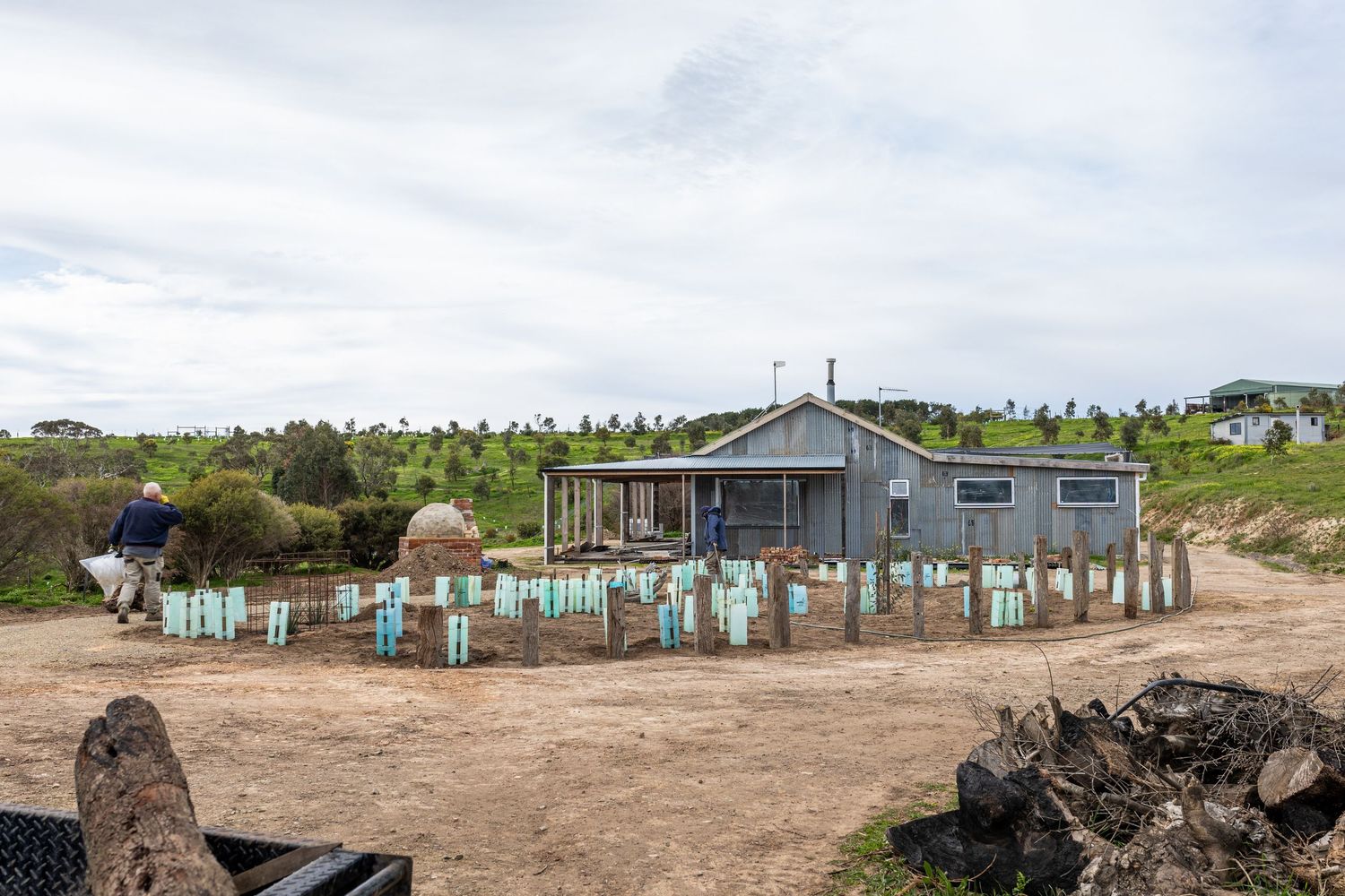 A rustic gray building with blue plant markers on a coastal hillside under cloudy skies in New Zealand.