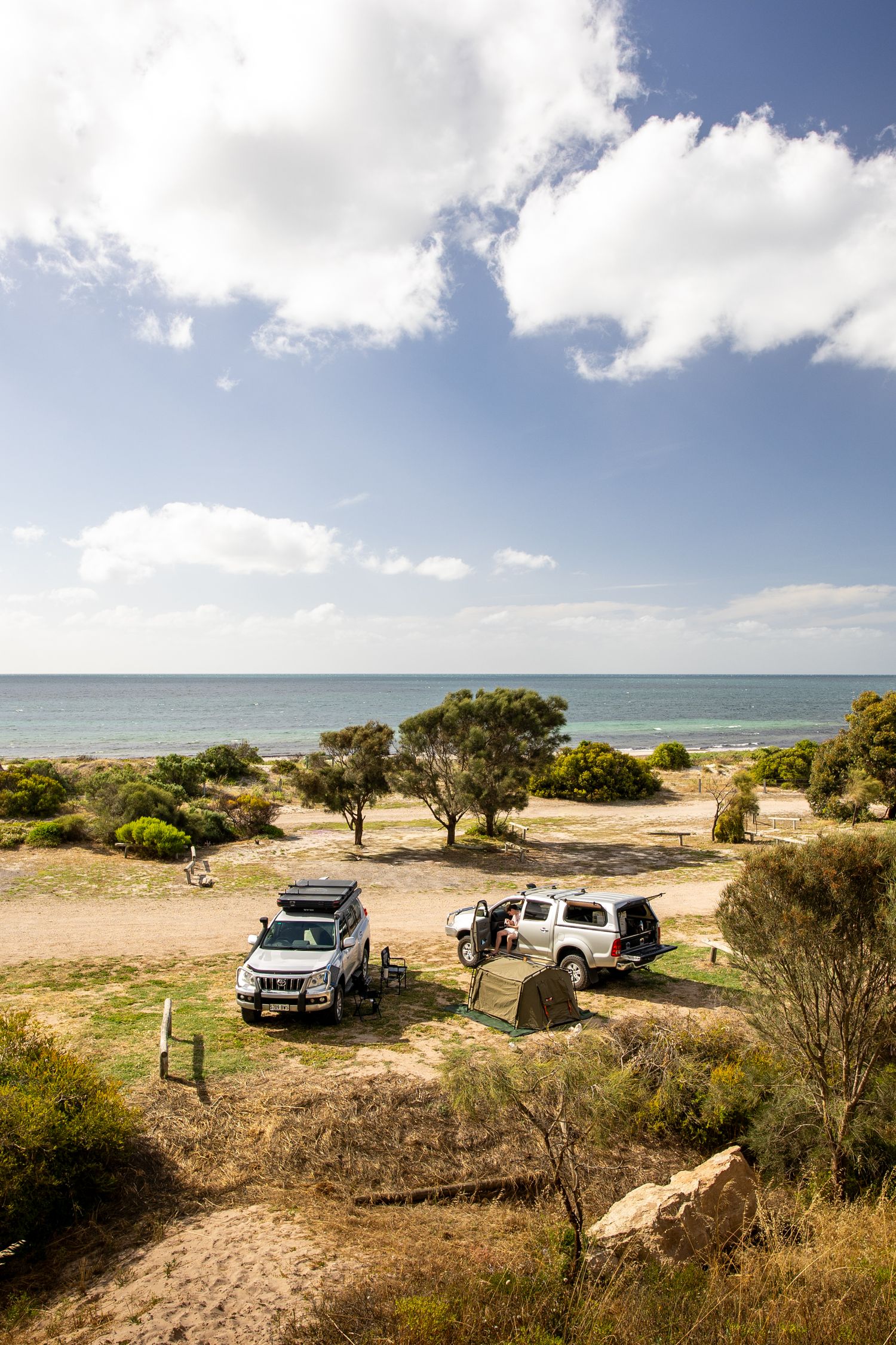 Two 4WD vehicles camped near the ocean with a tent set up on a sandy coastal campsite under a partly cloudy sky.