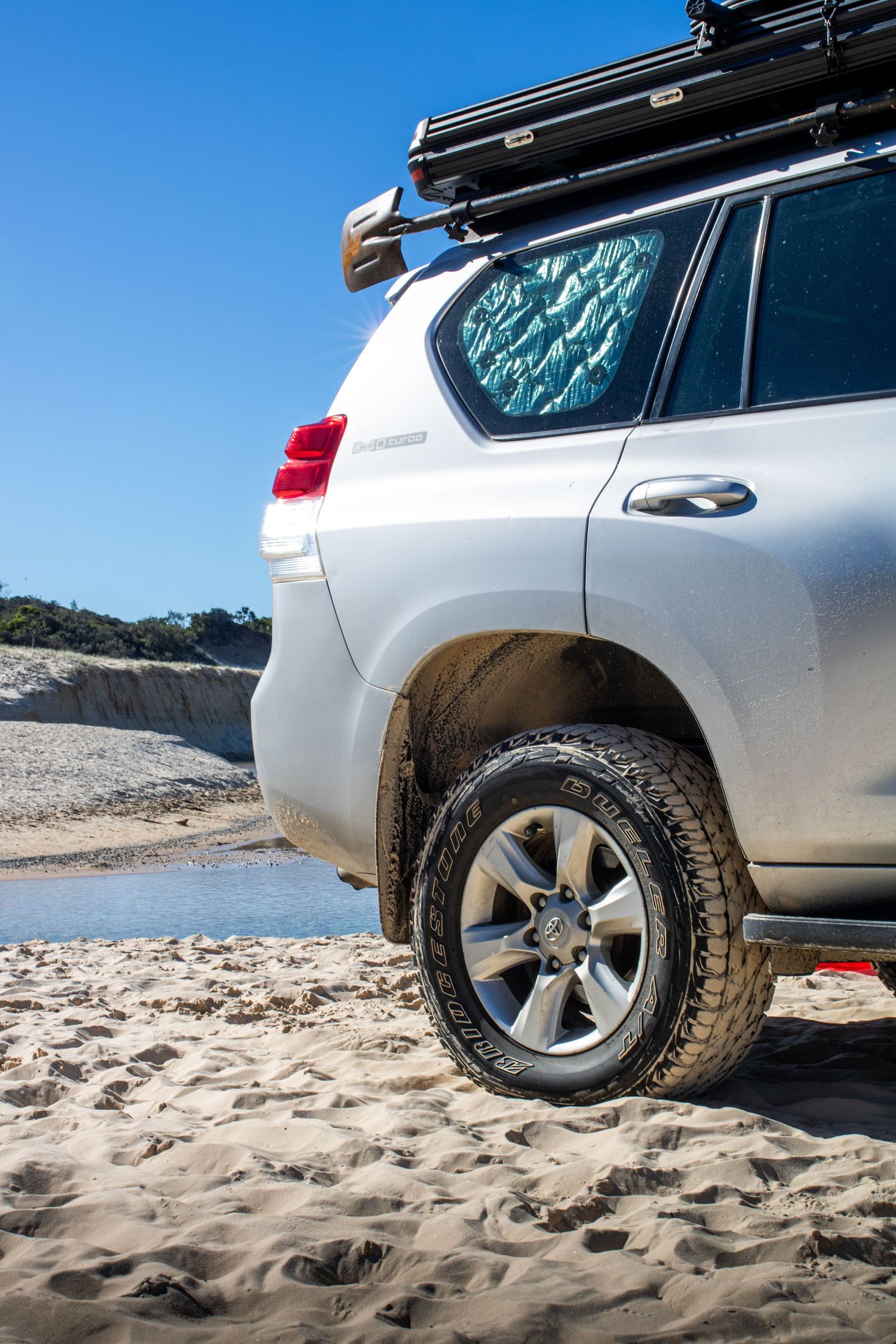 White off-road SUV with roof rack parked on sandy beach, showing muddy rear tire and blue sky backdrop.