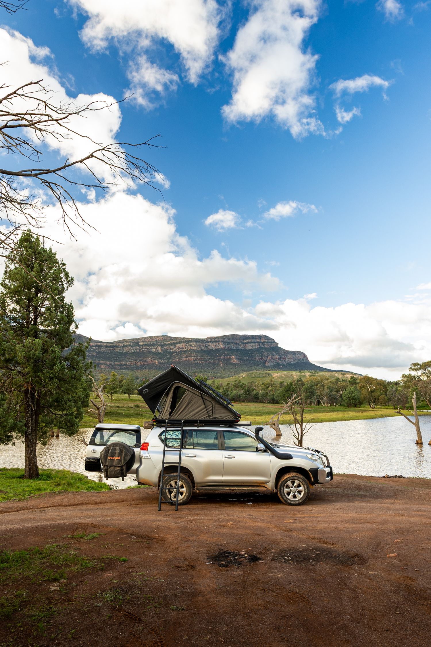4WD Toyota Land Cruiser with rooftop tent parked beside a serene lake with mesa cliffs and blue sky in Australia.