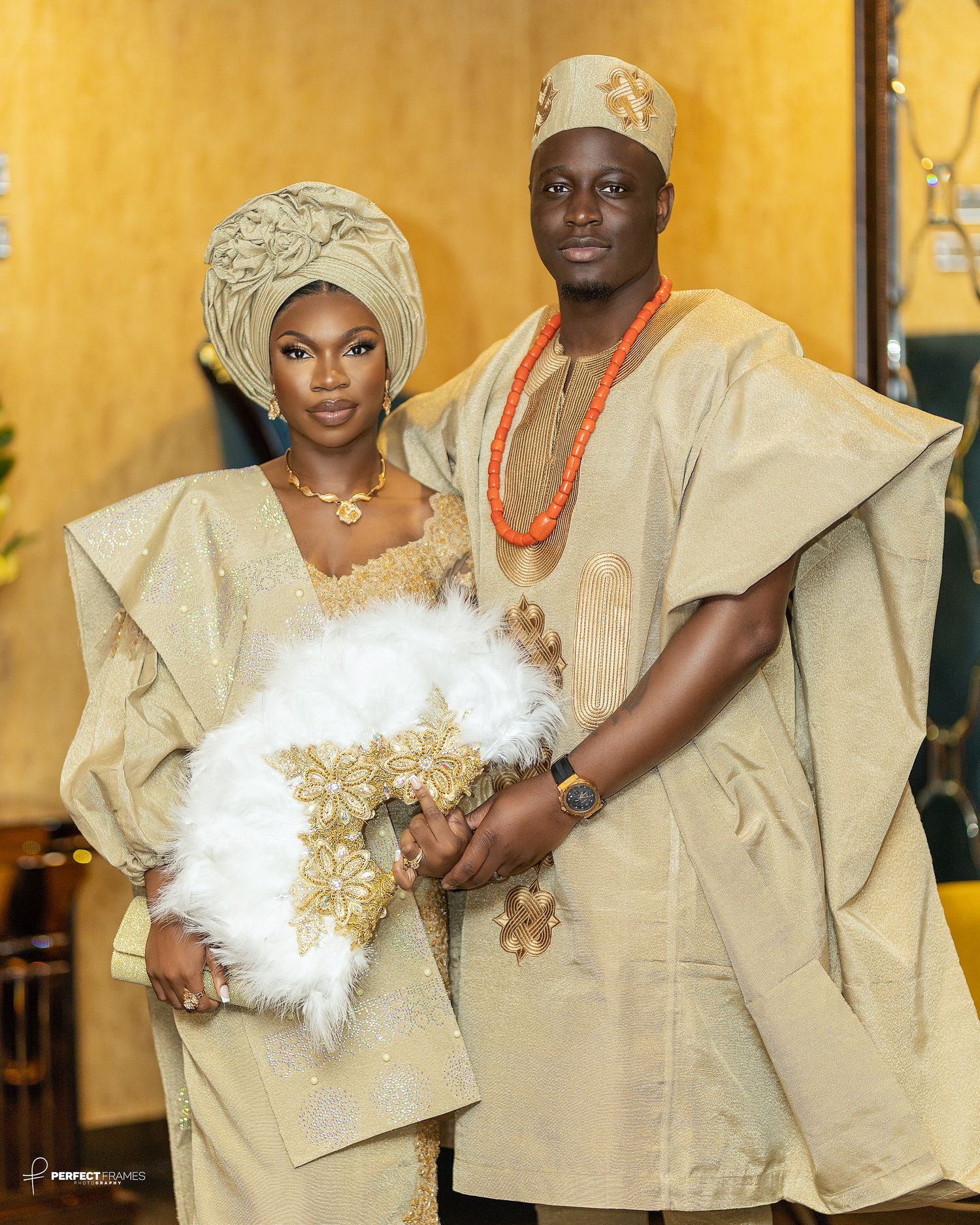 Nigerian traditional wedding couple in beige aso-oke attire, bride with gele, groom with beads, holding fan, cultural love celebration.