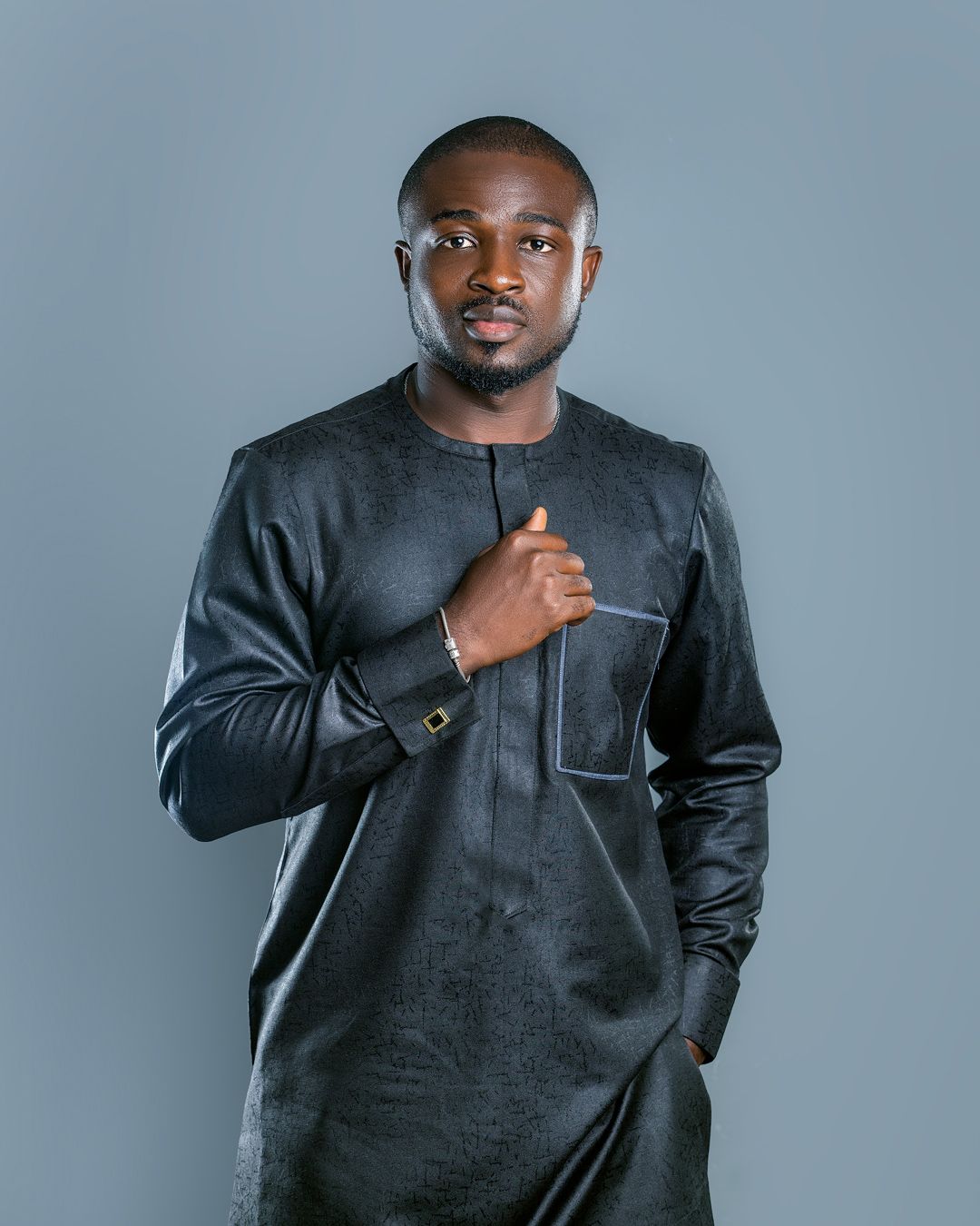Portrait of a confident African man in black traditional attire, hand on chest, studio lighting, dark background, modern cultural fashion.