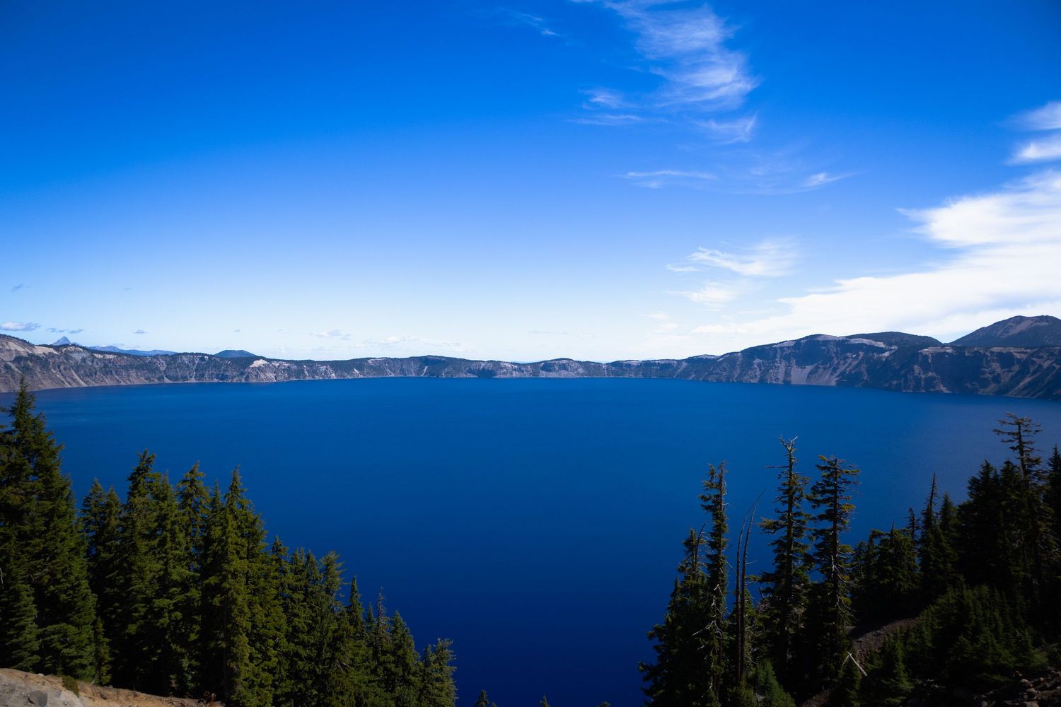 Panoramic view of Crater Lake's deep blue waters surrounded by steep cliffs and evergreen forests under clear skies.