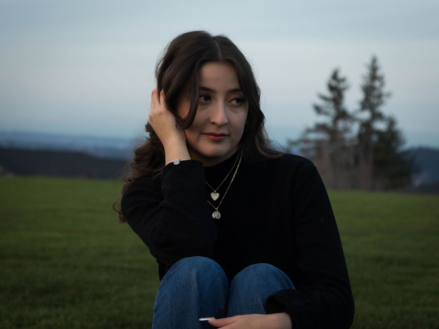 Someone in a black sweater and jeans sits outdoors against a scenic mountain landscape at dusk.