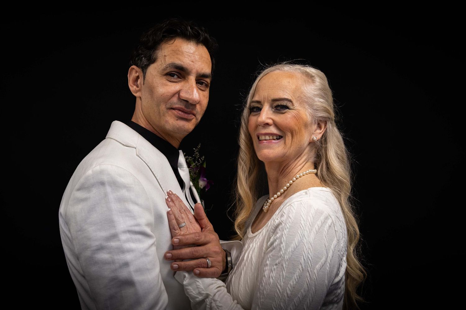 Elegant couple in white formal attire pose together against dark background during romantic photoshoot.