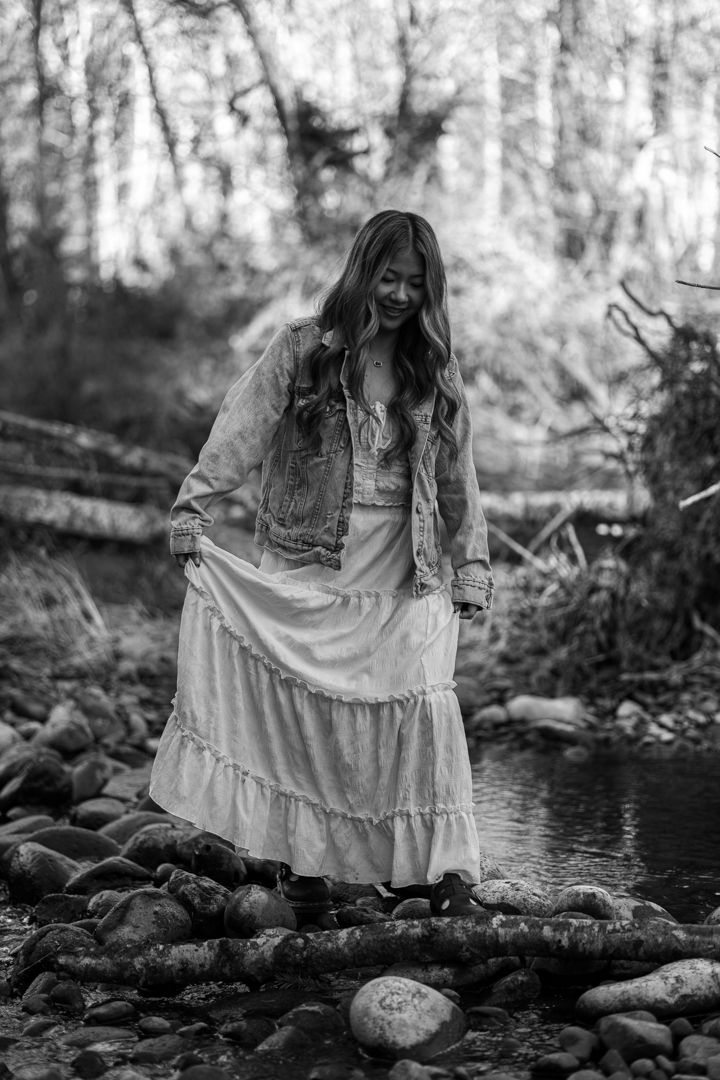 Black and white photo of a woman in a white maxi dress and denim jacket stepping on rocks by a forest stream.