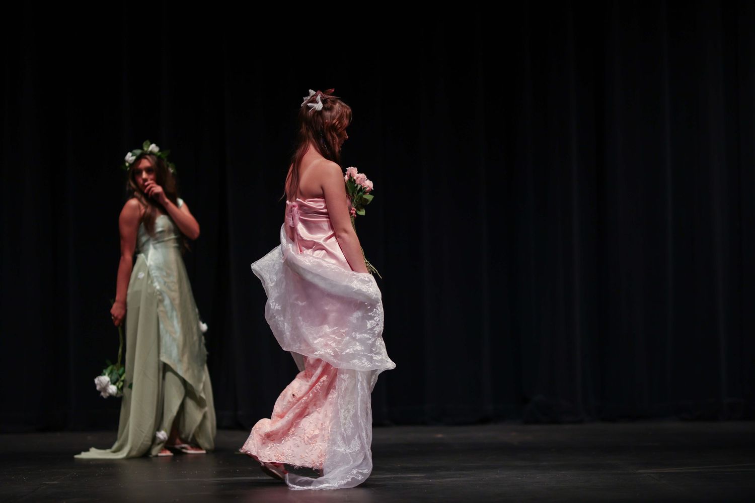Two ballet dancers in pink and olive dresses perform a graceful duet on a dark stage with dramatic lighting.
