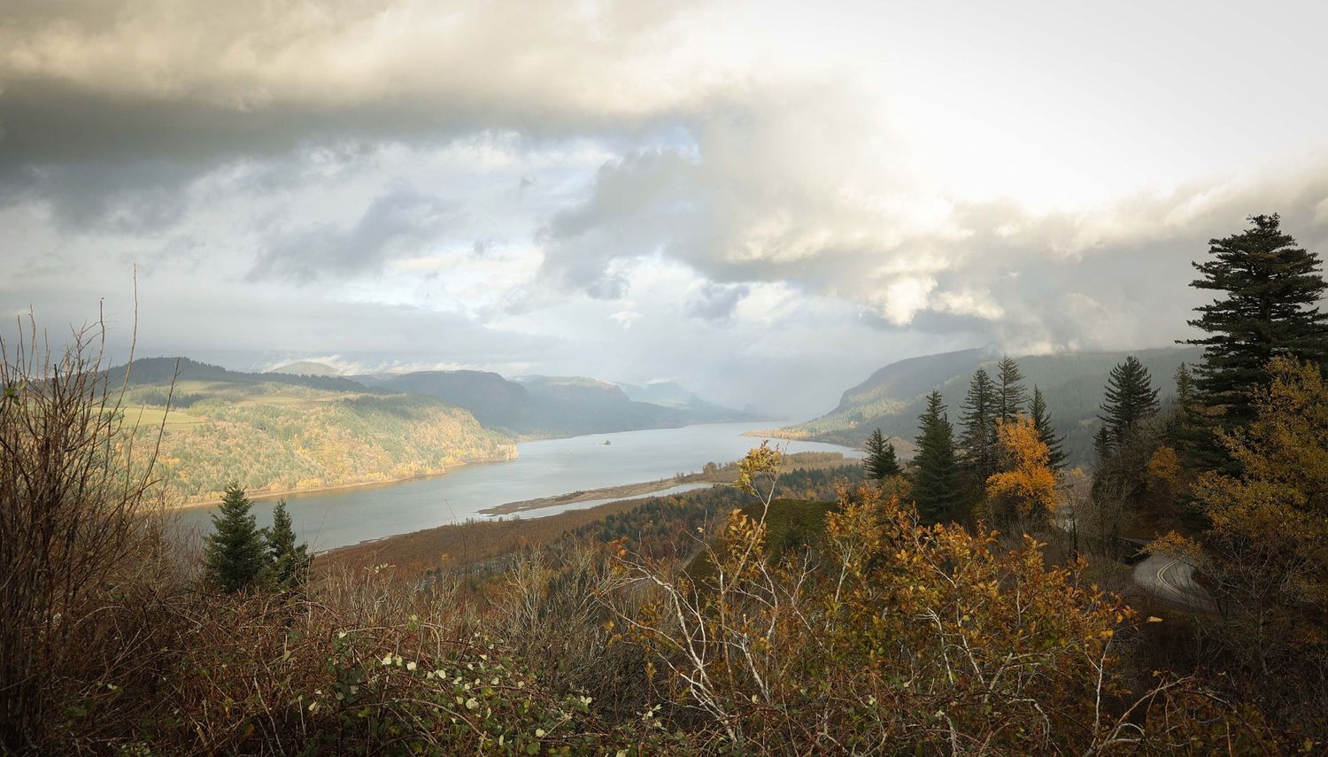 A panoramic mountain landscape with dramatic cloudy skies and evergreen trees along the ridgeline at sunset.
