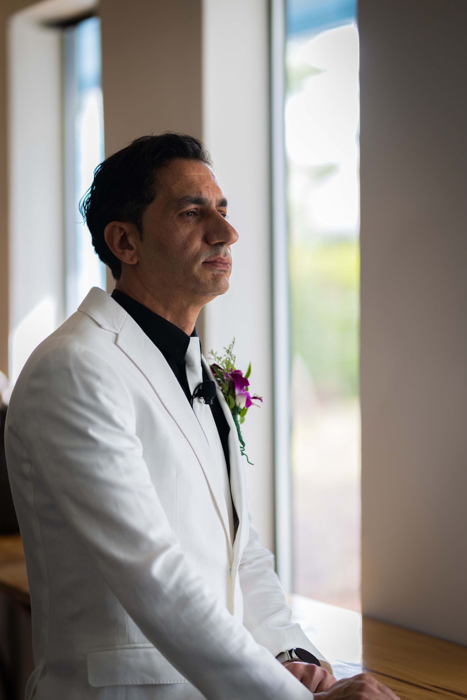 A person in a white tuxedo jacket with a boutonniere gazes thoughtfully out of a window in a contemplative pose.